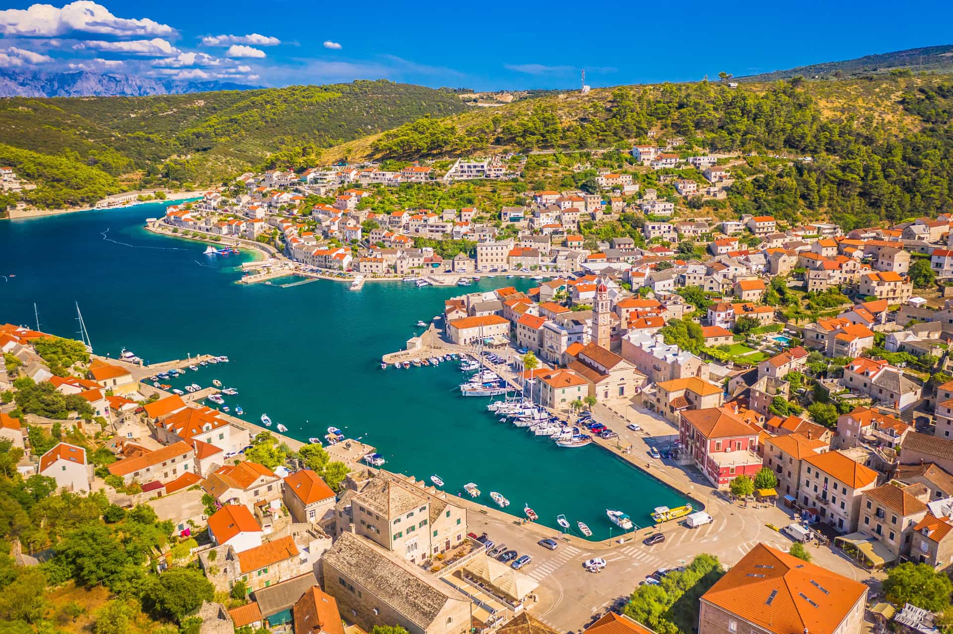 A beautiful seaside view of the coastal town of Pučišća on the island of Brač, Croatia—a popular small-ship cruise port destination. The image showcases the town's famous white limestone buildings with terracotta roofs terraced along a deep, protective bay of clear blue water, with several boats docked along the waterfront promenade under a bright sky.
