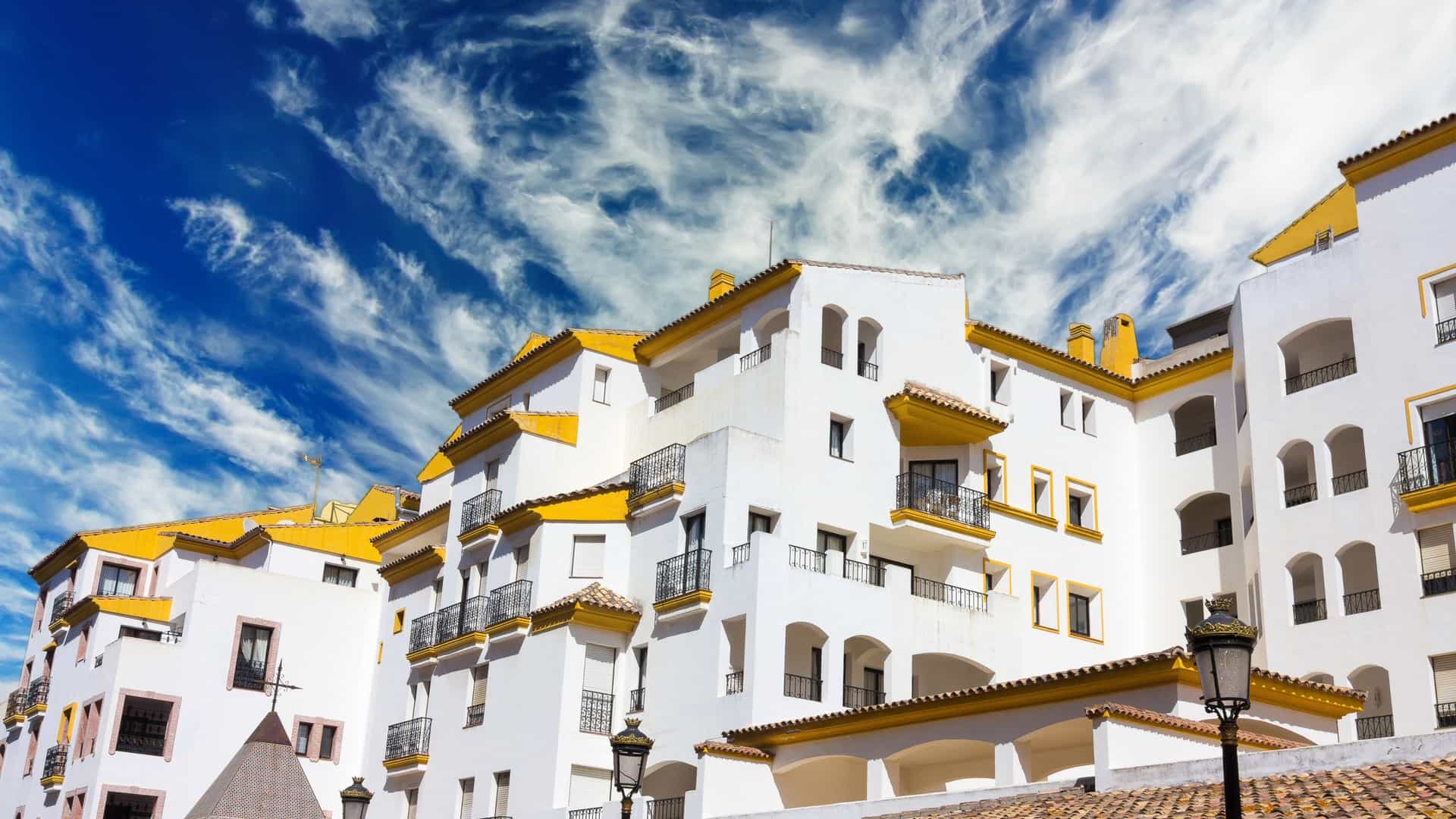 A high-angle scenic view of the luxury marina at Puerto Banús in Marbella, Spain—a glamorous Mediterranean cruise destination along the Costa del Sol. The image showcases rows of high-end yachts and luxury boats docked in the calm harbor waters, bordered by upscale white-washed buildings, boutique shops, and palm trees under a bright blue sky.