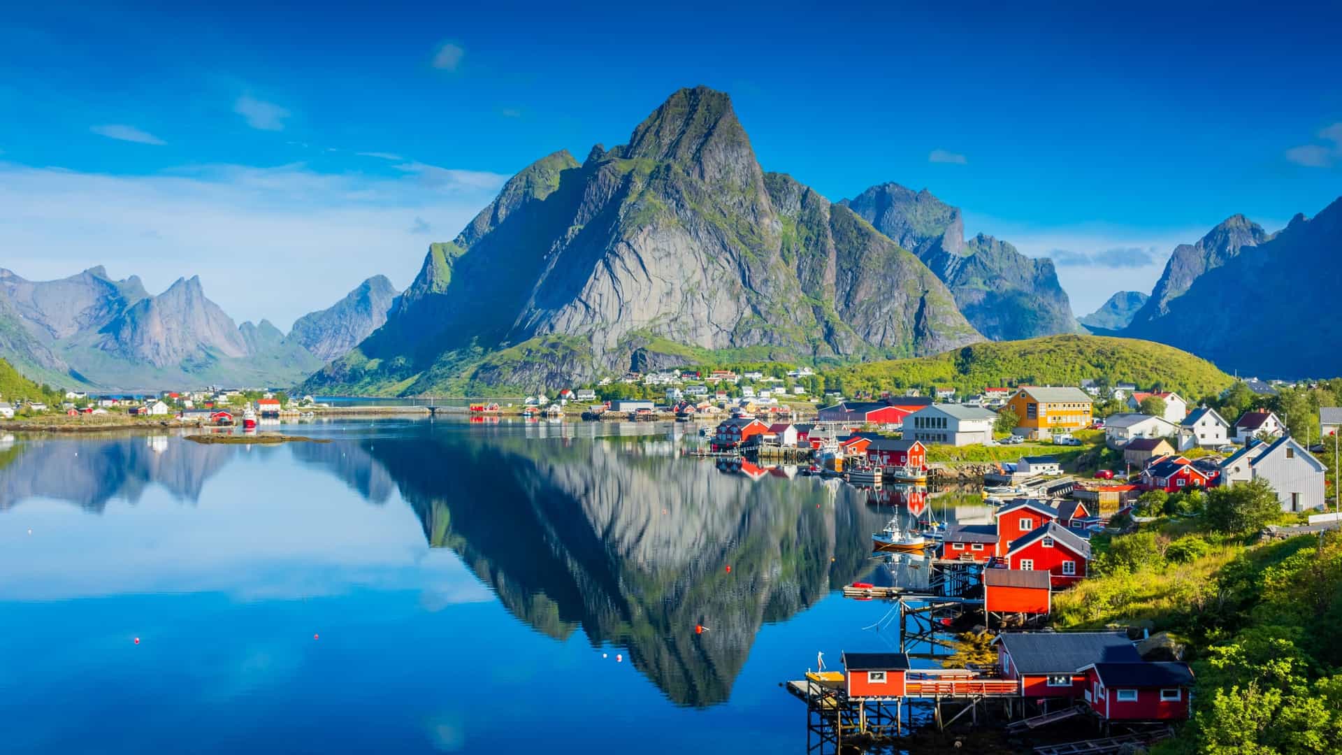 A breathtaking landscape view of the scenic fishing village of Reine, located on the Moskenesøya island in the Lofoten archipelago, Norway—a premier Arctic cruise destination. The image captures iconic red wooden fishermen's cabins (rorbuer) lining the calm waterfront, reflecting onto the glass-like sea, with the massive, snow-dusted peak of Reinebringen rising dramatically into a clear blue sky.