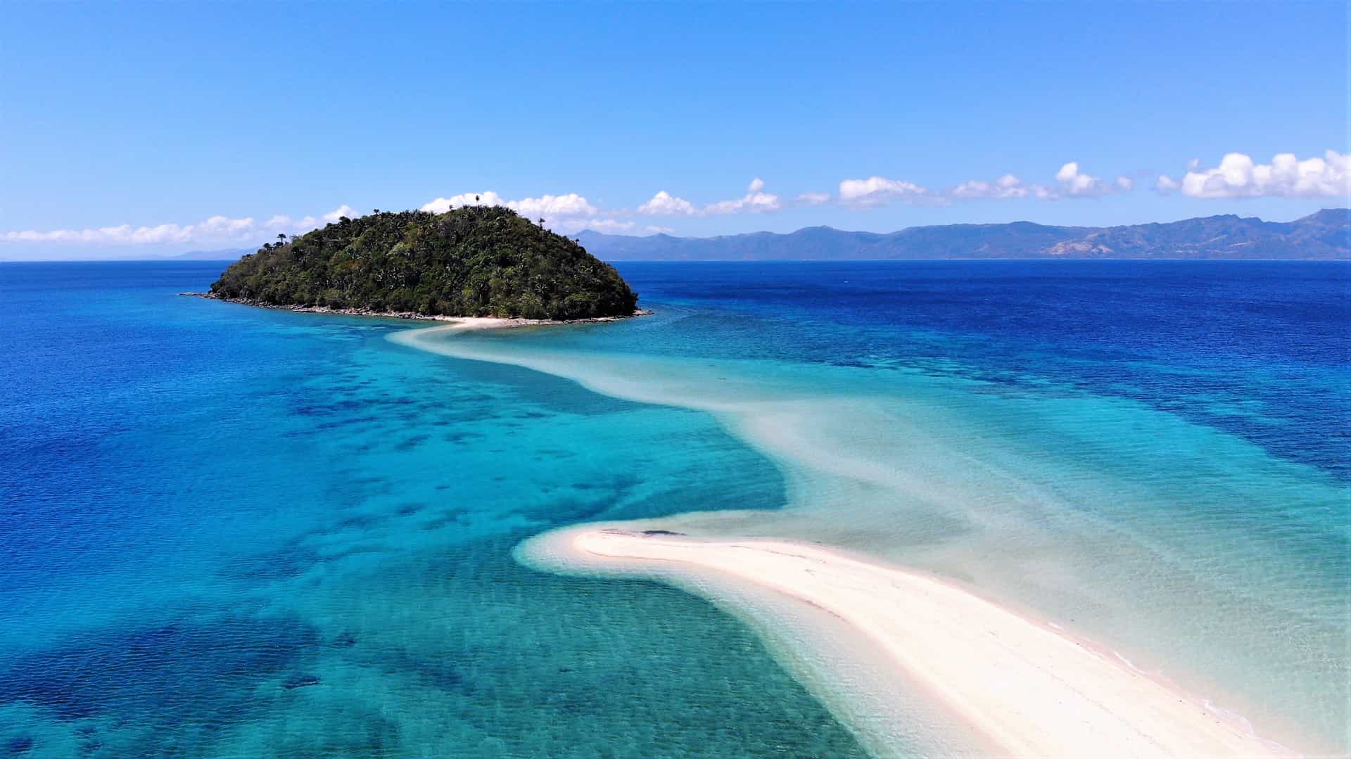 A spectacular aerial view of Bonbon Beach on Romblon Island, the Philippines—a pristine tropical cruise destination known for its natural beauty. The image captures a dramatic, curving white sandbar stretching out across crystal-clear turquoise waters toward a small, uninhabited islet covered in dense green jungle, with the deep blue sea and distant hills fading under a bright blue sky.