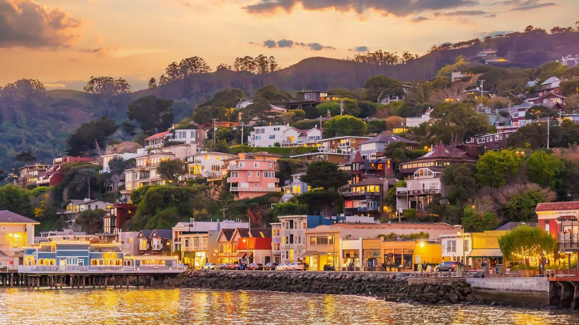 A beautiful twilight view of the hillside town of Sausalito, California—a popular ferry and shore excursion destination located just across the bay from the San Francisco cruise port. The image highlights charming, multi-tiered houses and waterfront restaurants terraced along the steep hills, with their lights reflecting onto the calm water under a warm golden sunset sky.