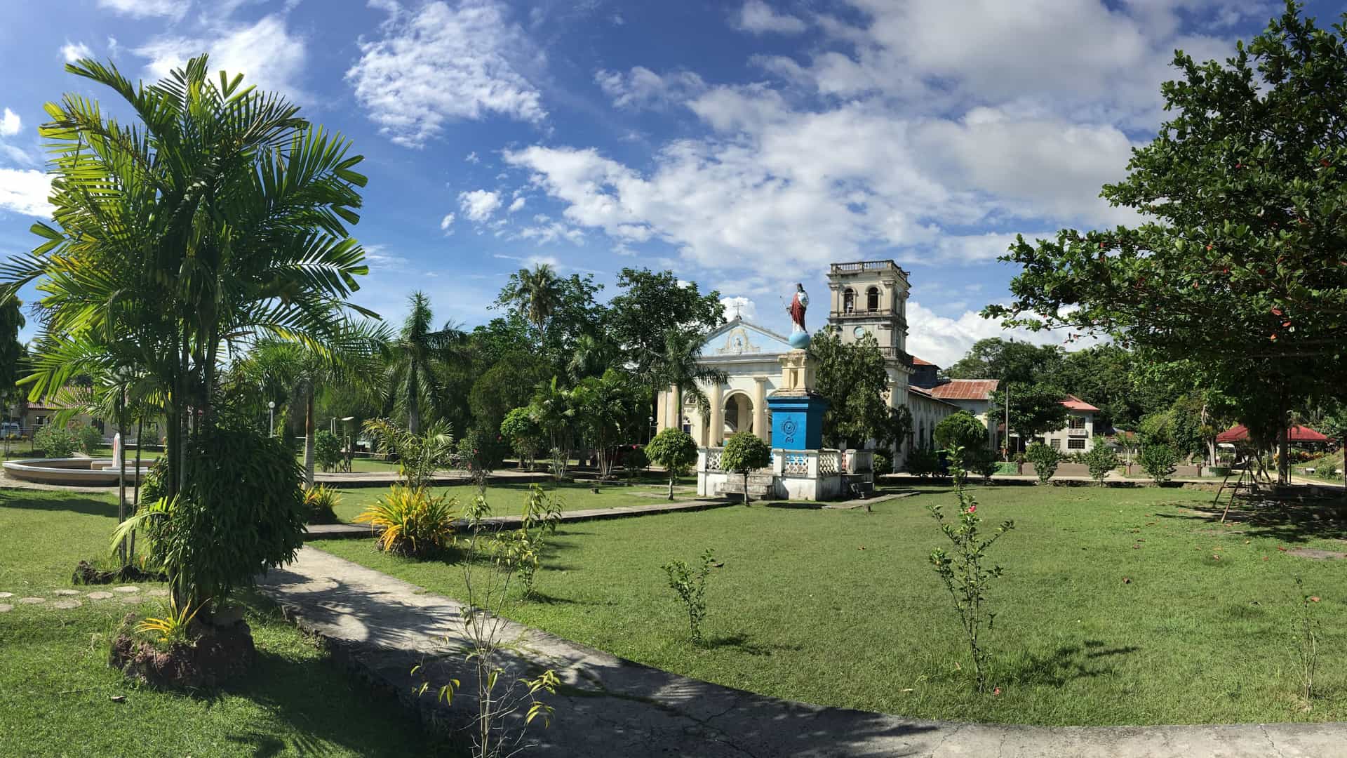 A sunny park scene in Tagbilaran, Bohol, featuring a historic church with blue accents, a religious statue on a pedestal, palm trees, and landscaped walkways that highlight the cultural charm of this Visayan cruise port.