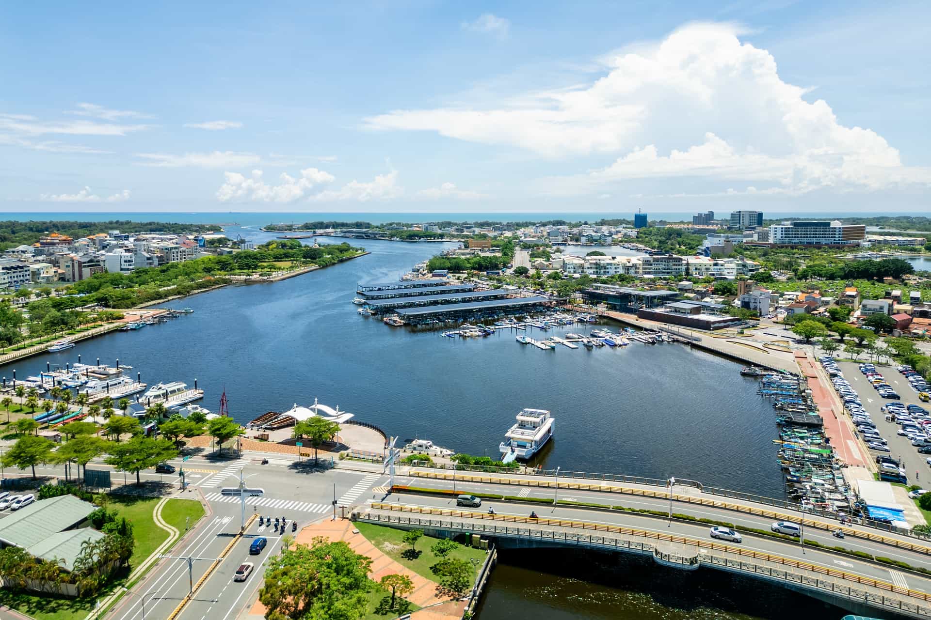 An aerial panorama of Anping Harbor in Tainan, Taiwan—a historic and cultural cruise port destination. The image captures the wide blue harbor waterway filled with modern covered boat docks, luxury yachts, and small vessels, bordered by coastal roads, green parks, seaside buildings, and a distant view of the open ocean under a bright, cloud-dotted sky.