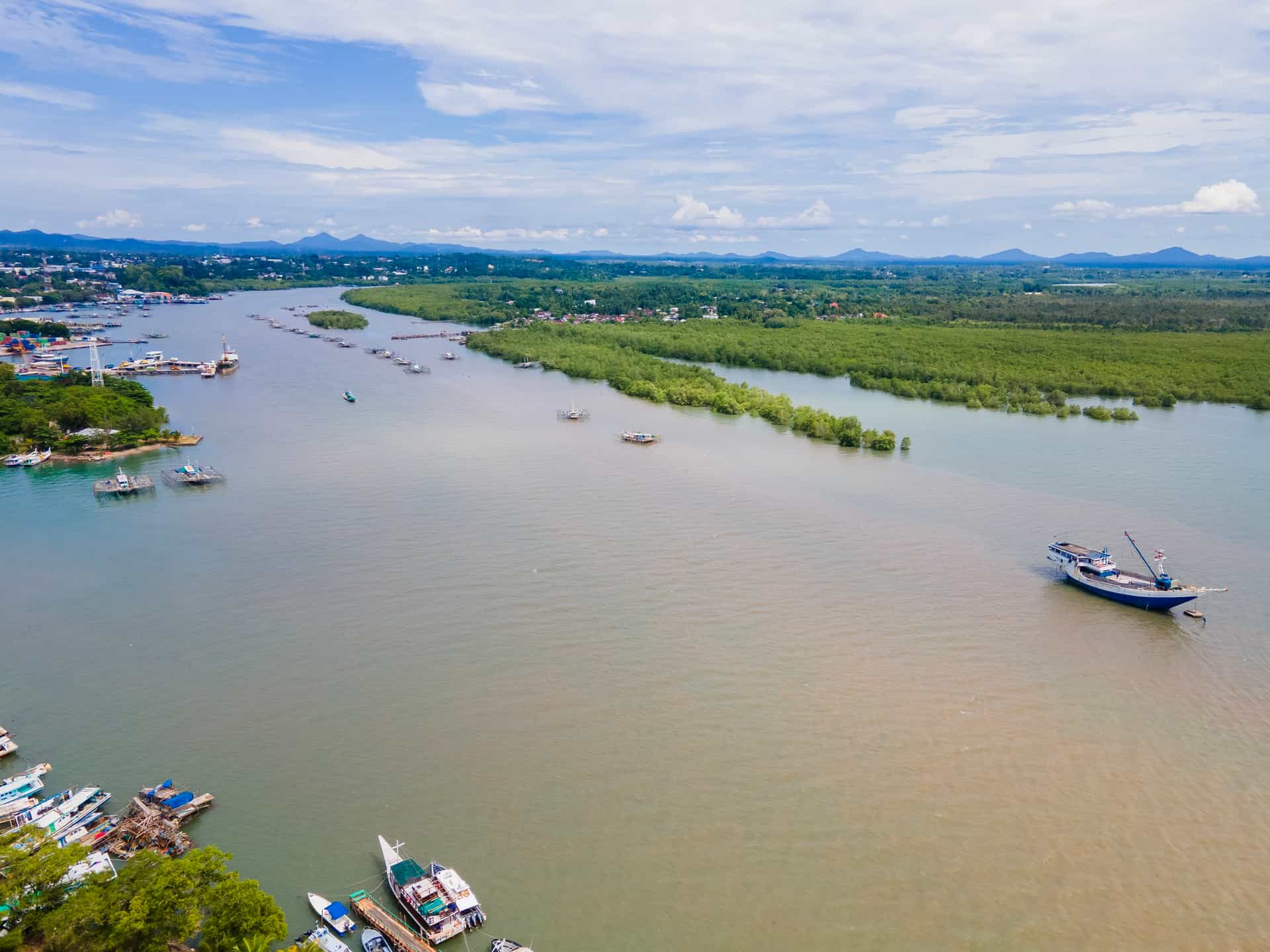 An aerial view of the Cerruc River estuary near Tanjung Pandan on Belitung Island, Indonesia—a unique Southeast Asian cruise stop. The image displays a wide, calm river channel where traditional wooden cargo ships and small fishing boats navigate, bordered on one side by a busy local port facility and on the other by dense, vibrant green mangrove forests stretching toward rolling hills in the background.