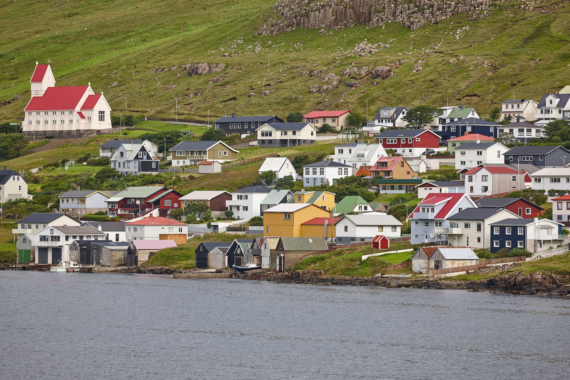 A stunning aerial view of the deep, blue fjord surrounding Tvøroyri on Suðuroy Island, part of the Faroe Islands (Kingdom of Denmark)—a dramatic and remote North Atlantic cruise destination. The image captures steep, rugged green volcanic mountains sloping sharply into the calm sea, with a few scattered buildings of the coastal village nestled along the shoreline under a vast, bright blue sky with wispy clouds.