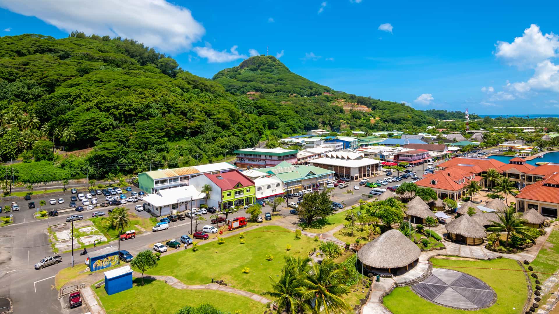 A sunny aerial view of Uturoa, Raiatea, showing colorful rooftops, tropical greenery, and forested hills rising behind the coastal town, capturing the vibrant landscape of this French Polynesia cruise port.