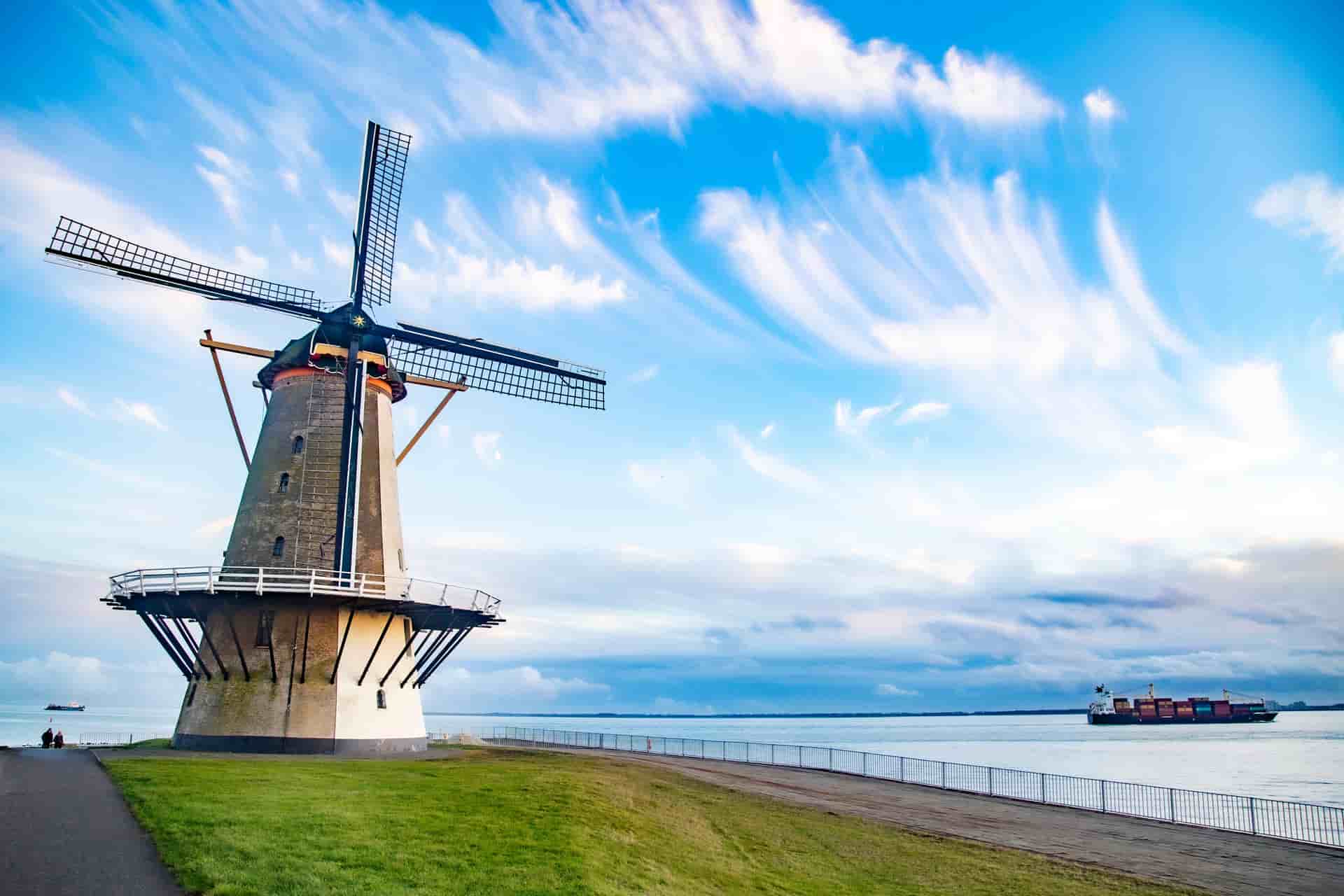 A striking waterfront view of the historic Oranjemolen (Orange Mill) windmill standing along the shoreline in Vlissingen, the Netherlands—a prominent Dutch maritime port. The image captures the traditional stone windmill on a grassy bank next to a coastal promenade, with a large container cargo ship passing by on the wide Scheldt river under a bright blue sky with dramatic, wispy clouds.