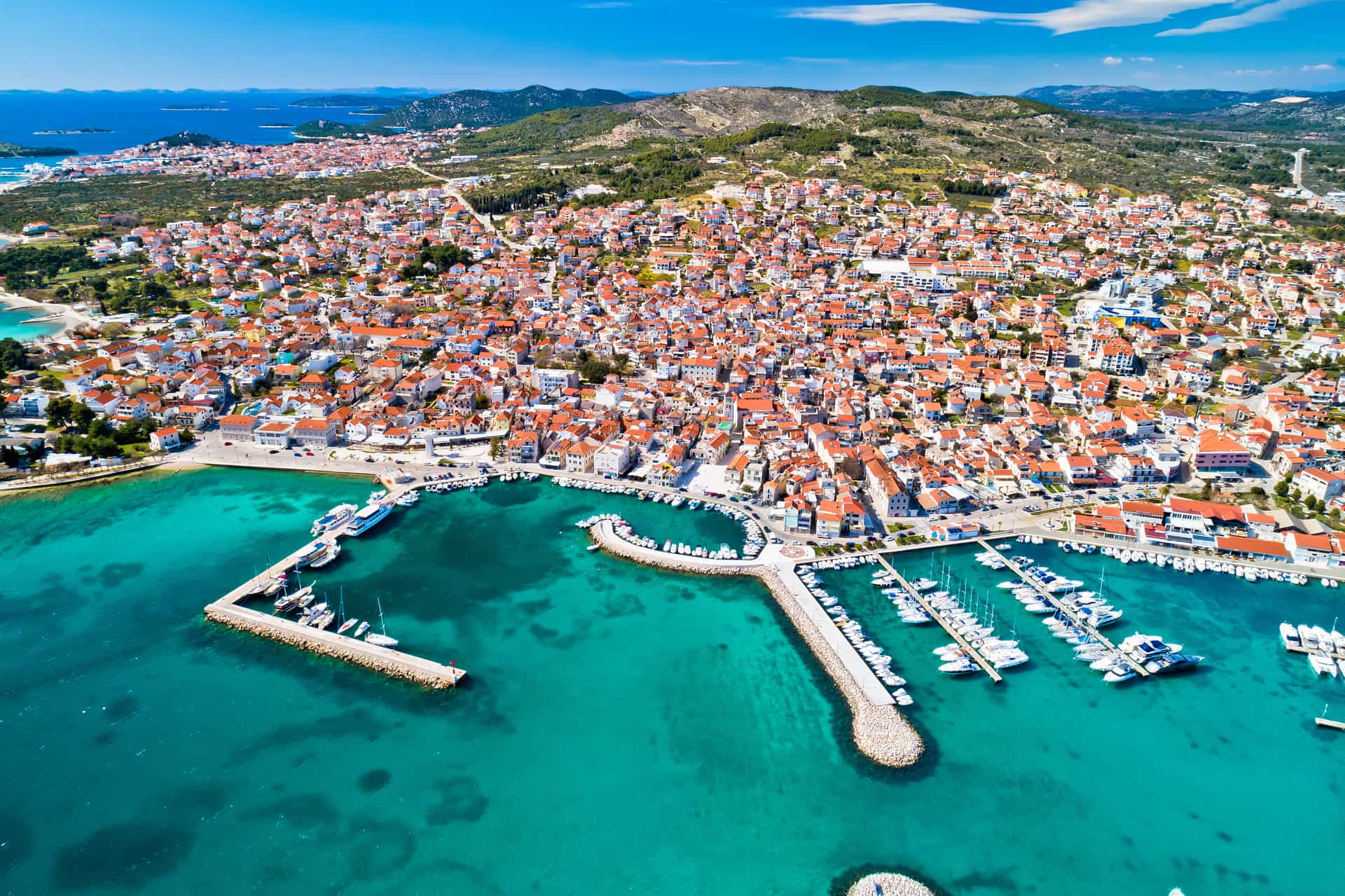 A high-angle aerial view of the coastal city of Vodice, Croatia—a vibrant port destination along the Adriatic Sea. The image showcases a sprawling town of densely packed stone buildings with orange terracotta roofs, wrapped around a large, protective marina harbor filled with small boats, docks, and piers jutting into crystal-clear turquoise and deep blue waters.