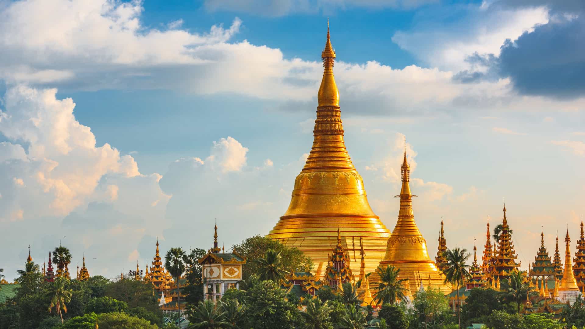 The golden Shwedagon Pagoda rising above lush greenery in Yangon, Myanmar, with ornate stupas and dramatic sunlit clouds highlighting this iconic Southeast Asian cultural landmark.