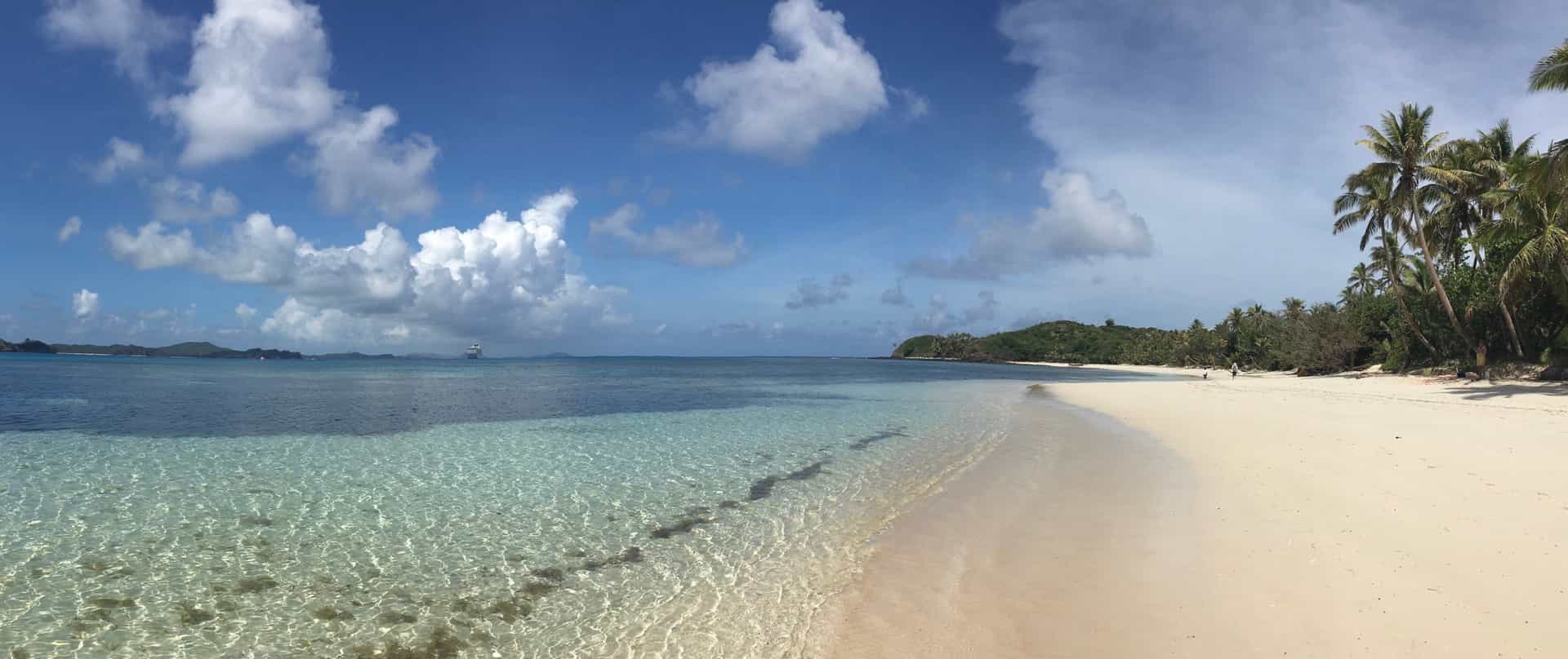 A beautiful shoreline view of the expansive white sand beach at Yasawa-i-Rara on Yasawa Island, Fiji—a pristine South Pacific cruise destination. The image shows gentle, transparent turquoise waves lapping against the shore, bordered by a dense canopy of leaning palm trees and lush green vegetation stretching toward a low green hill under a blue sky with scattered clouds.