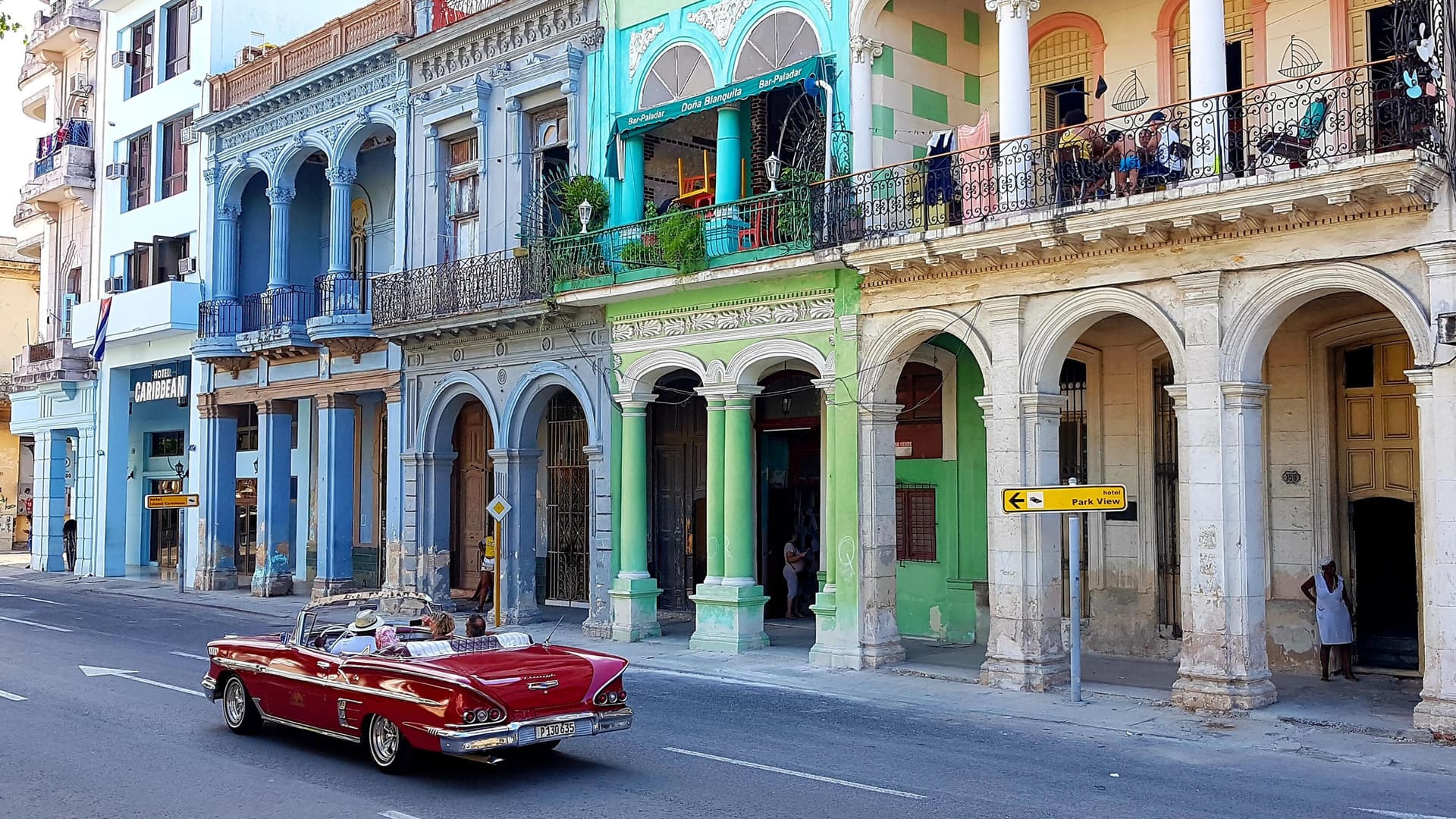 A vintage red convertible driving down a street lined with brightly painted buildings.