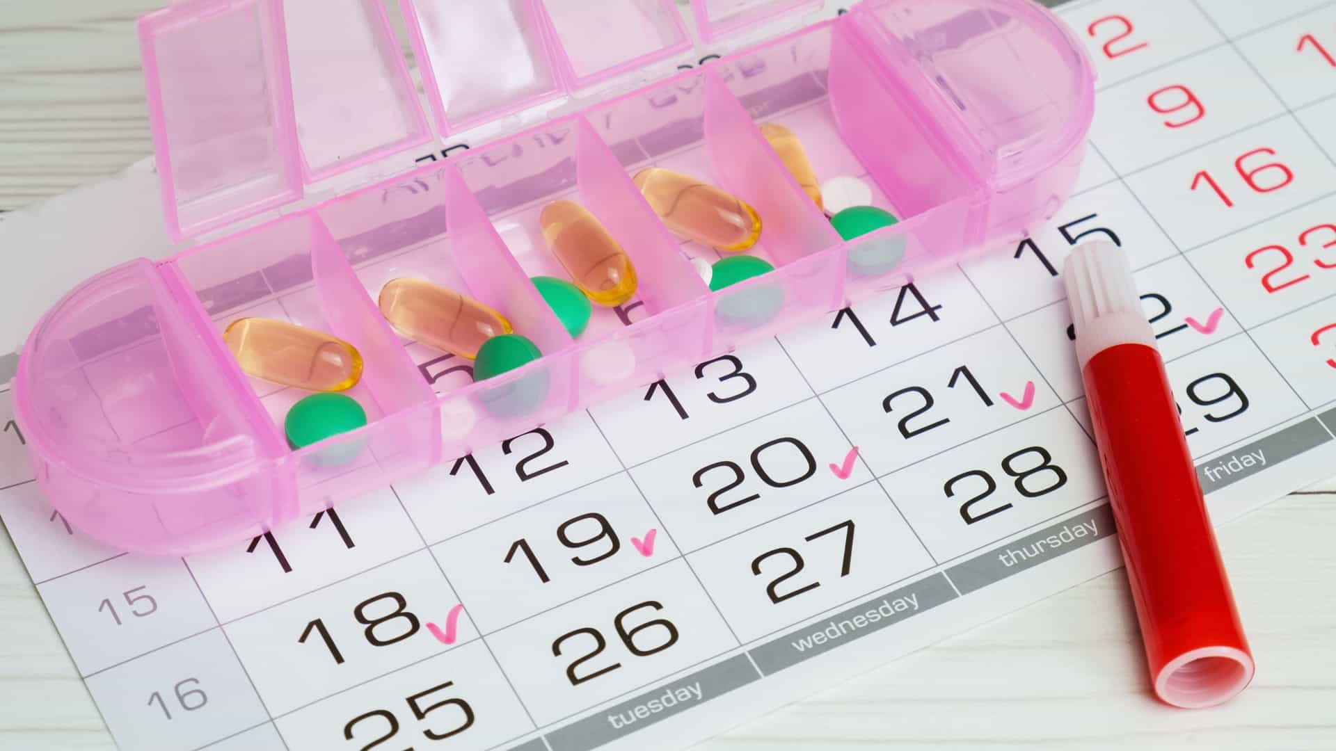A pink daily pill box for medication management laid on top of a calendar with days checked off.