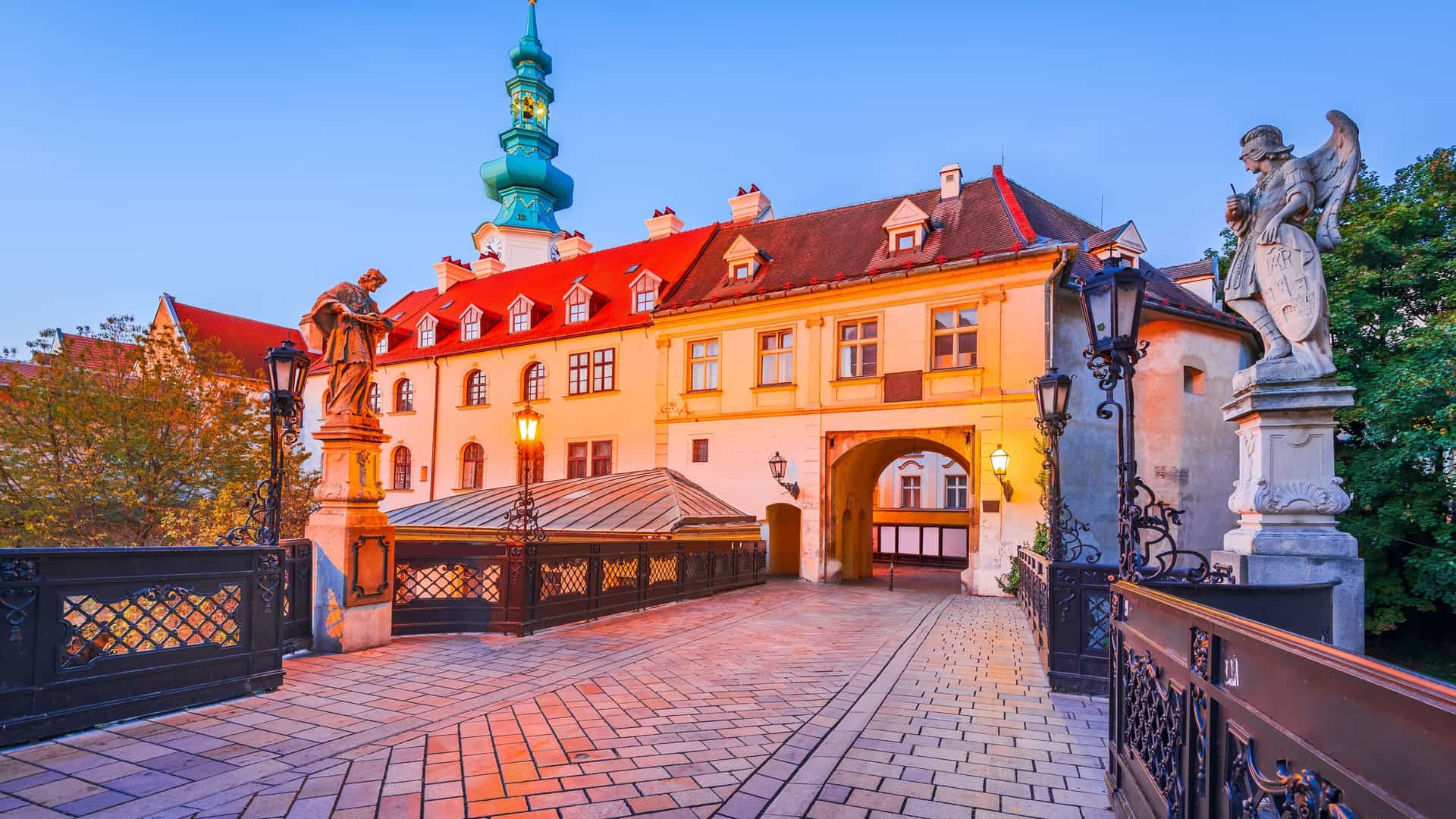 An evening view of St. Michael's Gate, a historic landmark in Bratislava's Old Town, Slovakia, located near the Danube River. The cobblestone bridge in the foreground is illuminated by old-fashioned street lamps and leads to the gate's archway. The main building has a distinctive red-tiled roof and white facade. To the left, a tall, green-domed tower with a clock rises against the twilight sky. The image beautifully captures the historical architecture and charming atmosphere of the city, which is a popular stop on Danube River cruises.