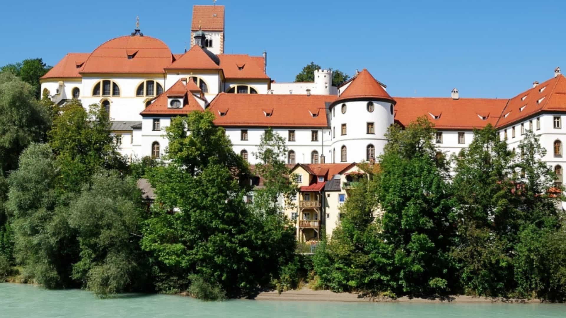 The picturesque Benedictine Abbey of Weltenburg in Germany, situated on the banks of the Danube River. The large, white-walled building with its distinctive red roofs, domes, and a prominent round tower rises above a line of green trees and the turquoise water of the river. The abbey, a famous stop on many Danube cruises, stands out against a clear, bright blue sky, showcasing its historical and architectural grandeur.