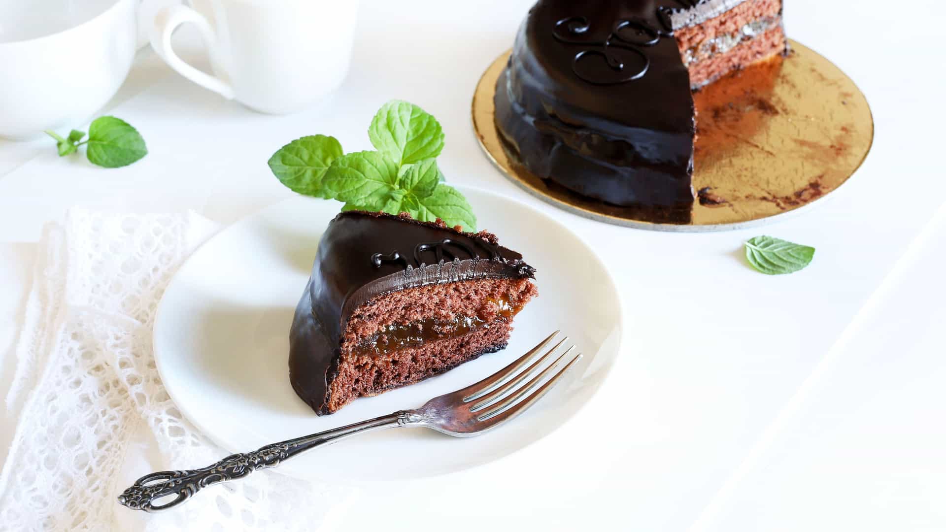 A delicious slice of Sachertorte, a famous Austrian chocolate cake, is presented on a white plate with a vintage-style fork. The slice reveals layers of dense chocolate sponge cake and a thin apricot jam filling, all covered in a rich, dark chocolate glaze. A sprig of fresh mint garnishes the plate. In the background, the rest of the round cake is visible, with one slice missing. This image is associated with Austria, a key destination along the Danube River.