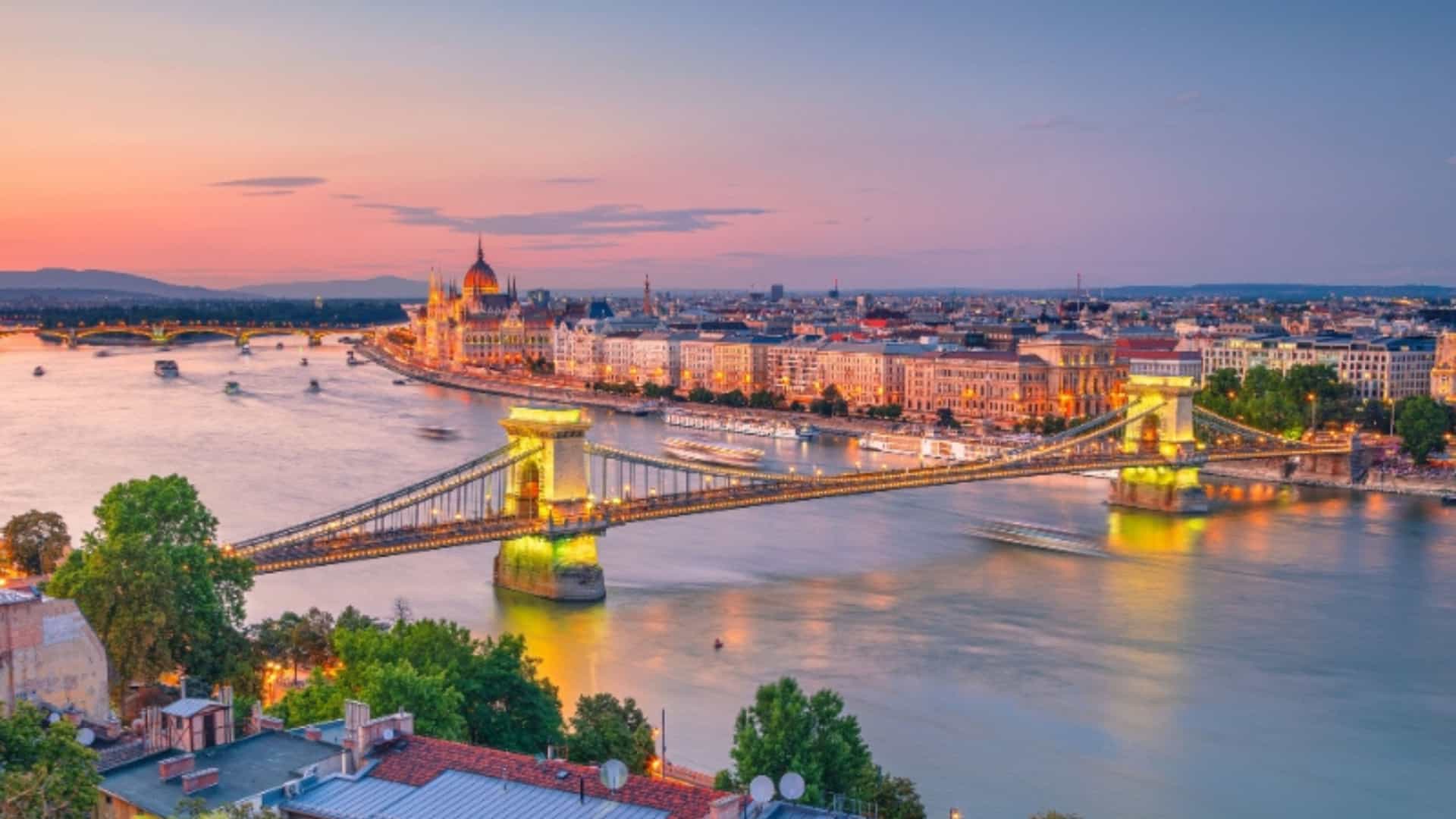 An iconic evening view of the Chain Bridge in Budapest, Hungary, spanning the Danube River. The bridge is illuminated with golden lights, contrasting with the twilight sky. On the far side of the river, the grand Hungarian Parliament Building is visible, also lit up, along with a line of brightly lit historic buildings along the riverbank. The river itself shows the blurred trails of boats, suggesting movement. The image beautifully captures the illuminated cityscape of Budapest, a key city along the Danube River.