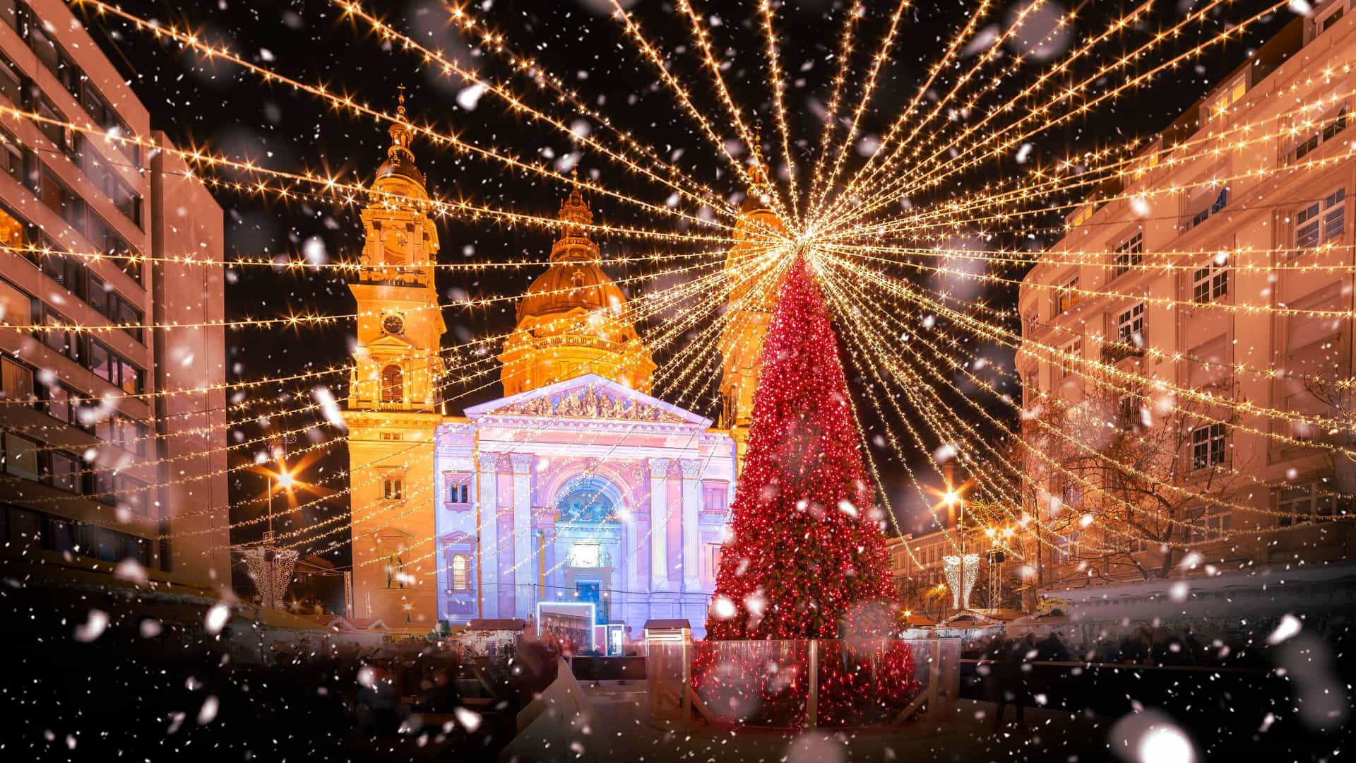 An enchanting nighttime view of St. Stephen's Basilica in Budapest, Hungary, during the Christmas season. The basilica's facade is illuminated with colorful lights, and a massive, brightly lit red Christmas tree stands in the square in front of it. A network of glowing string lights radiates out from the top of the tree, creating a festive canopy over the square. Snowflakes are falling, adding to the magical atmosphere. This scene captures the vibrant holiday spirit of a major city along the Danube River.