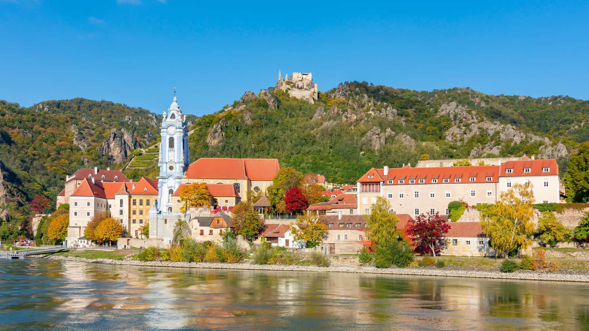 A picturesque view of Dürnstein, a small town on the banks of the Danube River in Austria. The town's prominent landmark, the Dürnstein Abbey with its blue and white church steeple, stands out. Behind it, a mix of red-roofed buildings and colorful trees of the Wachau Valley line the riverbank. High on the wooded hill in the background, the ruins of Dürnstein Castle are visible. The image, a classic scene from a Danube River cruise, captures the serene beauty of the Austrian countryside in autumn.