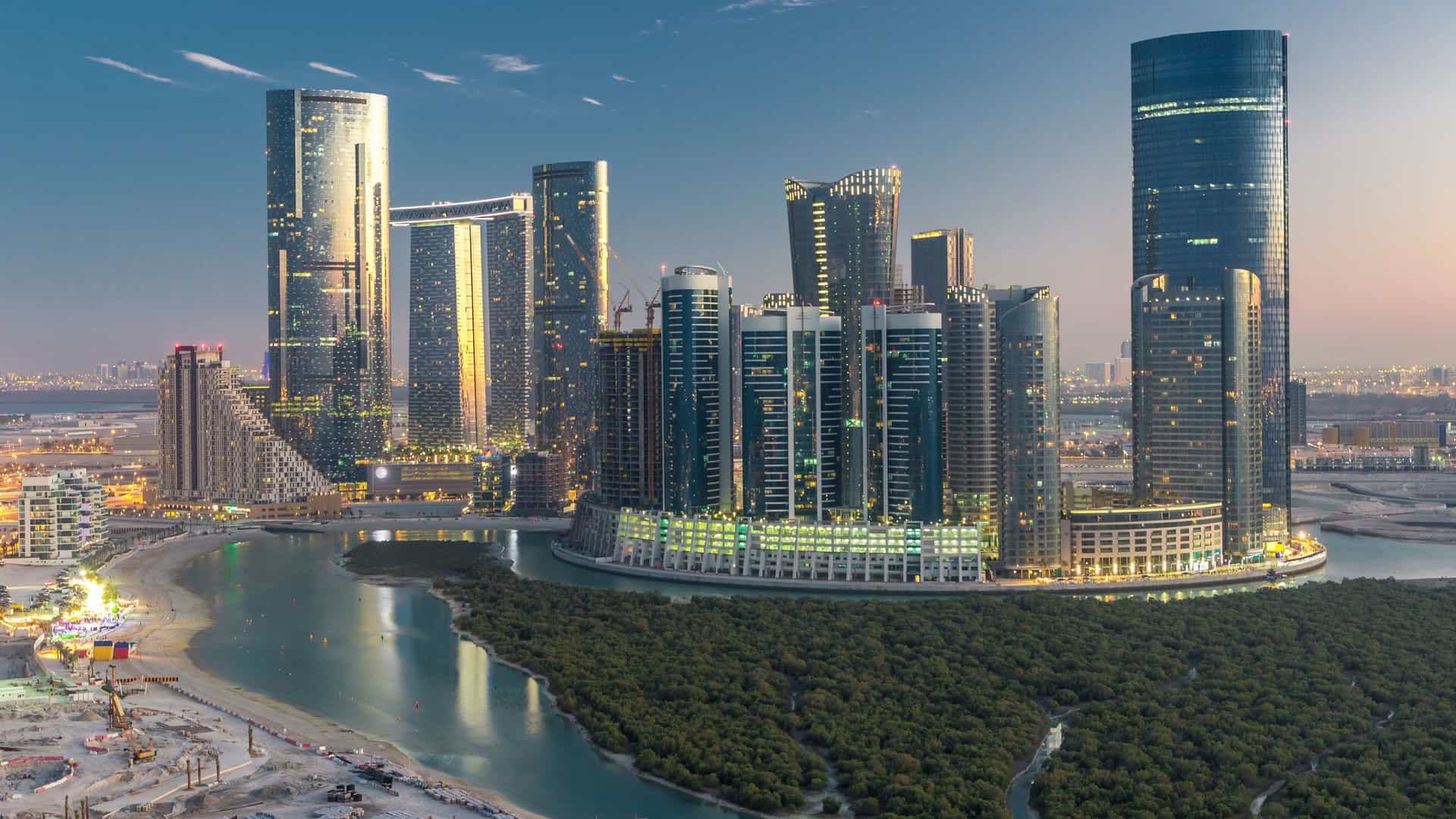 Aerial view of the modern skyline of Al Reem Island, Abu Dhabi, featuring illuminated glass skyscrapers and towers at twilight. The cityscape shows a day-to-night transition over the Persian Gulf.