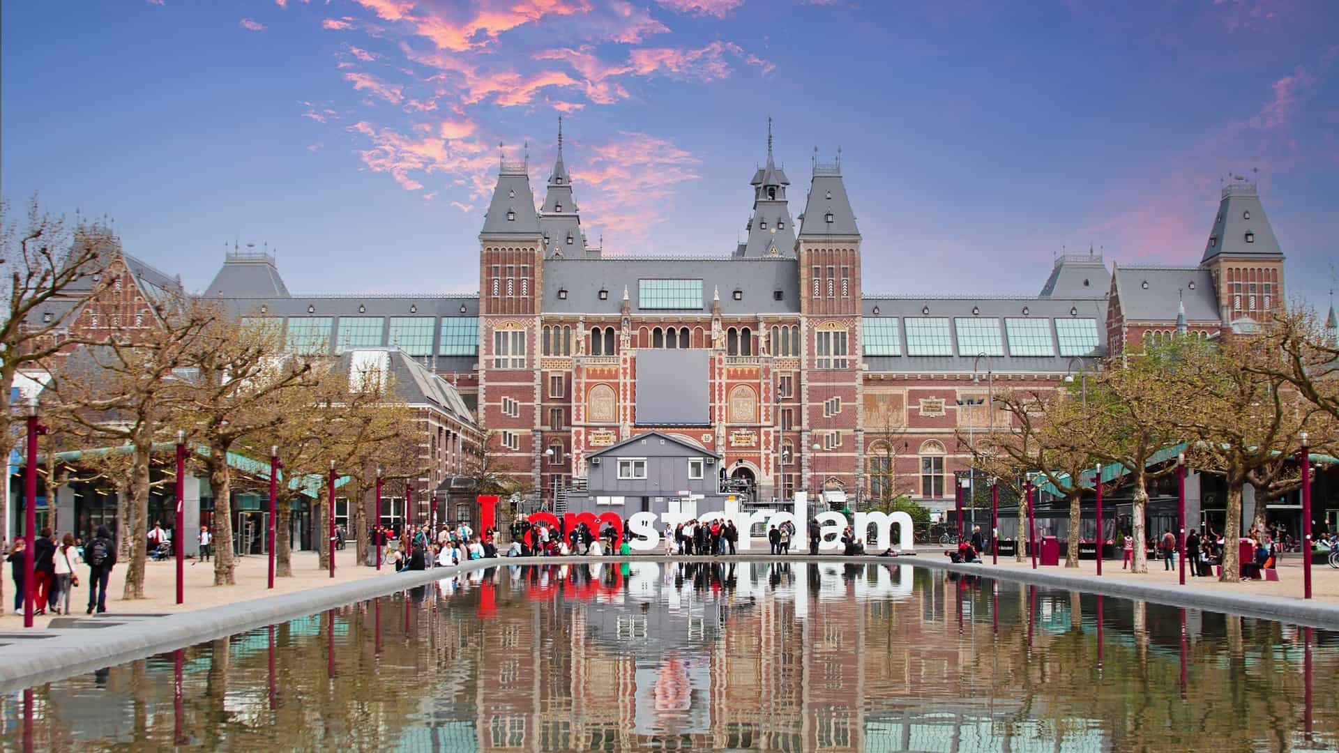 A view of the Rijksmuseum, the grand Dutch national museum in Amsterdam, Netherlands, photographed at sunset. The Neo-Gothic architectural facade, featuring red brick and elaborate ornamentation, is illuminated by the warm, golden light of the setting sun, possibly showing the famous archway passage with crowds or cyclists.