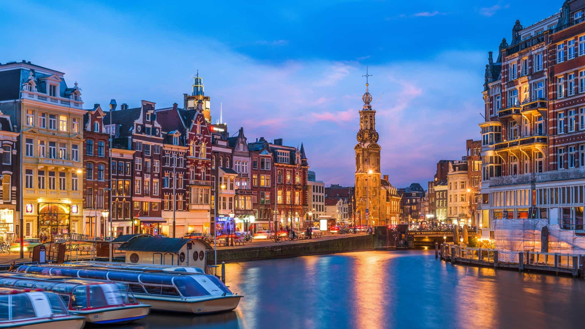 A scenic night shot of Amsterdam's canals at twilight, with the city's historic buildings and the illuminated Munttoren Tower reflecting brightly on the calm water, creating a stunning cityscape.