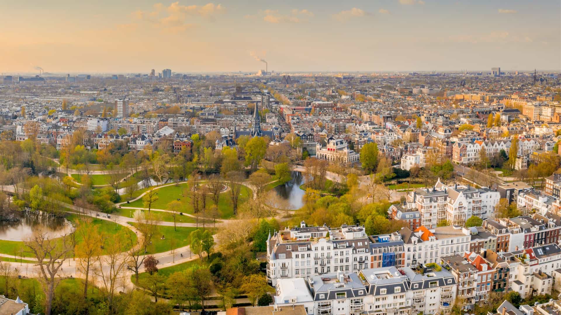 An aerial view of Vondelpark, a large public park in Amsterdam, Netherlands. The image shows a vast expanse of green trees and lawns, crisscrossed by paths, with a pond visible within the park's boundaries. The park is surrounded by the city's urban landscape.