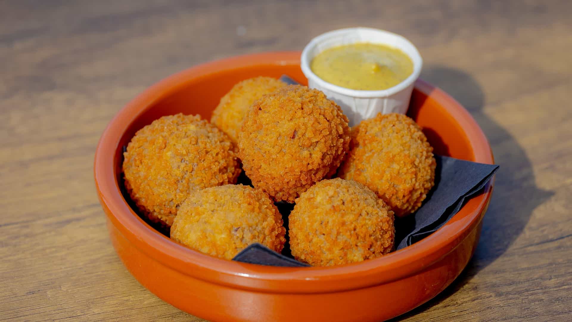 A close-up, high-angle view of several golden-brown, deep-fried Dutch bitterballen (meat-based croquettes) arranged on a white serving plate, accompanied by a small ramekin of yellow mustard for dipping. The plate is set against a wooden background.