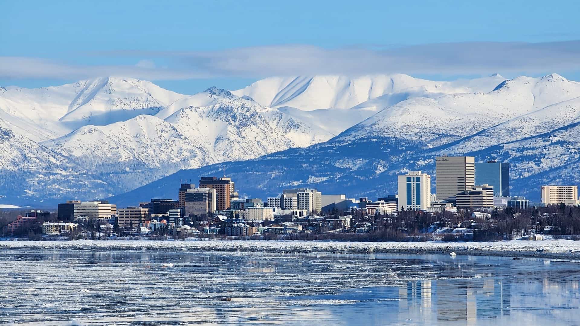 The skyline of downtown Anchorage, Alaska, viewed across a partially frozen inlet, with the Chugach Mountains rising behind it.