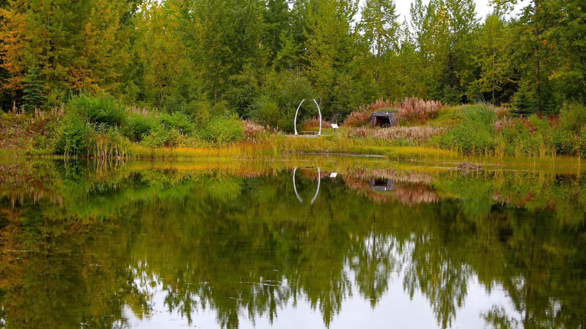A scenic view of the Alaska Native Heritage Center complex across the calm waters of Lake Tiulana. The main modern center building is visible in the distance, and the lake's shoreline features several traditional, life-sized Alaska Native village dwellings, surrounded by dense Alaskan forest.