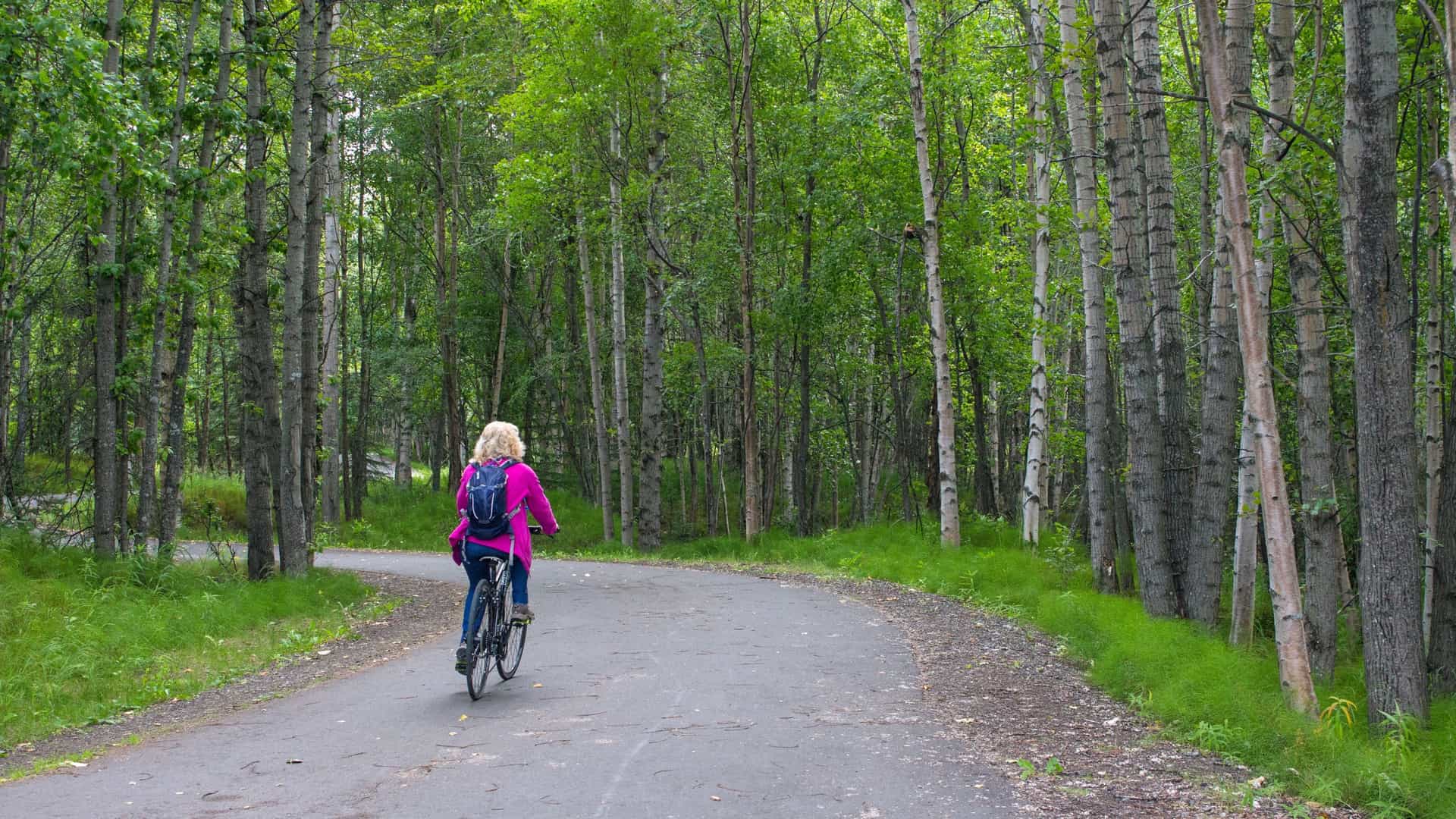 A person on a bicycle rides along the paved Tony Knowles Coastal Trail in Anchorage, Alaska. The trail is bordered by a lush, green forest on one side and opens to a scenic view of the Cook Inlet and surrounding mountains on the other.