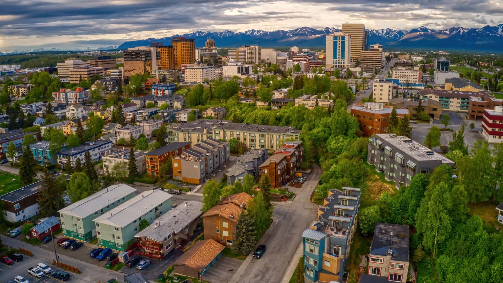 An aerial, panoramic view of the downtown Anchorage, Alaska skyline. Modern office buildings and city streets fill the foreground, while the background is dominated by the vast, snow-capped peaks of the Chugach Mountains and the waters of the Cook Inlet.