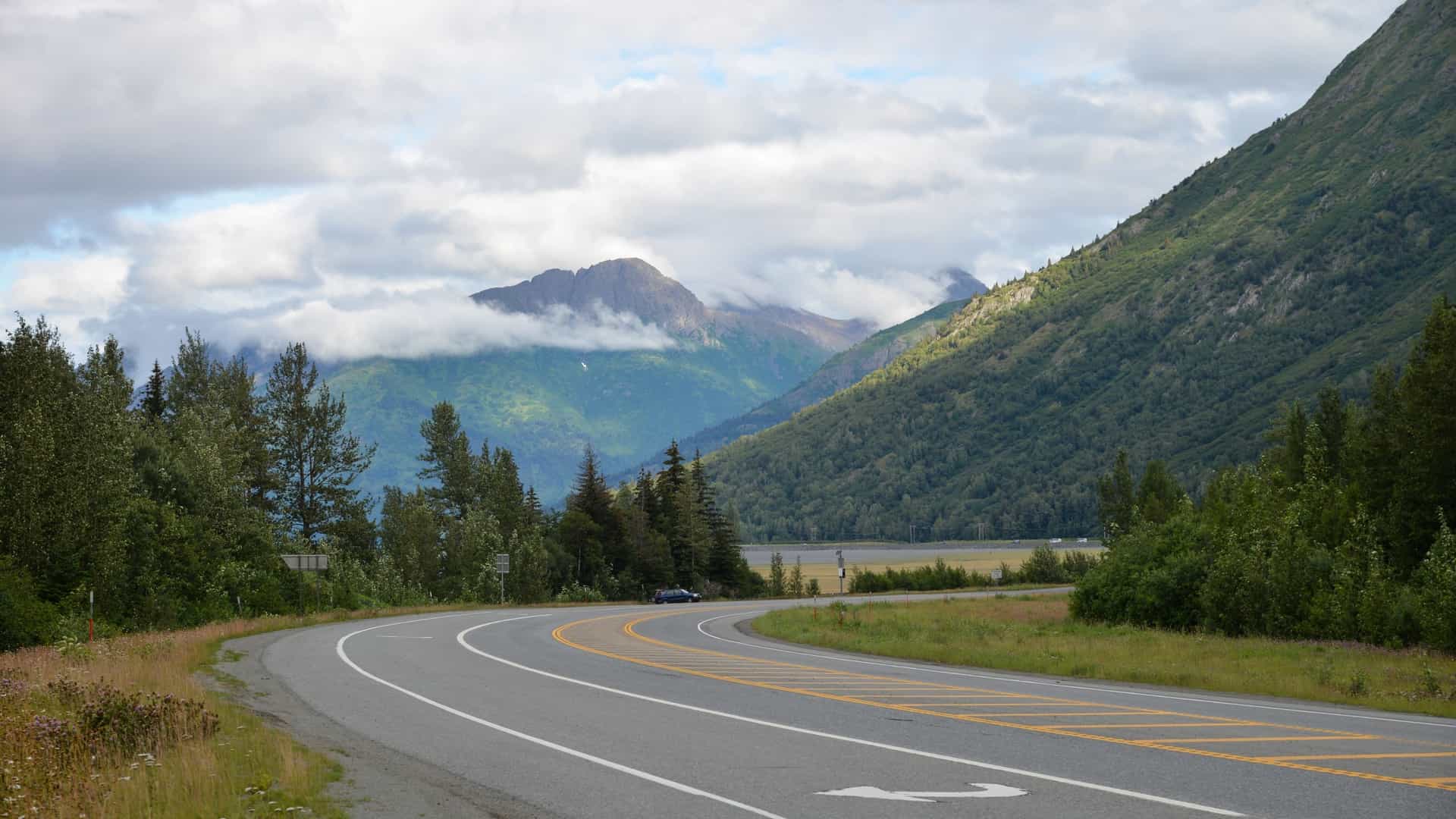 A dramatic scenic view along the Seward Highway overlooking Turnagain Arm in Alaska. The glacial silt-filled, muddy waters of the tidal inlet stretch into the distance, bordered by the steep, forested slopes of the Chugach Mountains, a common viewing spot for the bore tide.