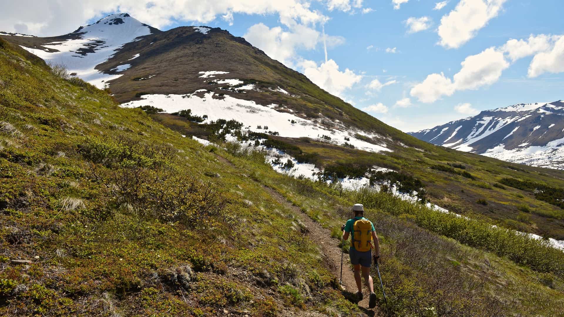 A rear-view, vertical photo of a female mountain climber walking uphill on a steep, rocky trail in Chugach State Park, Anchorage, Alaska. She is wearing a backpack and hiking gear. The trail is surrounded by rugged, mountainous terrain under a clear sky.