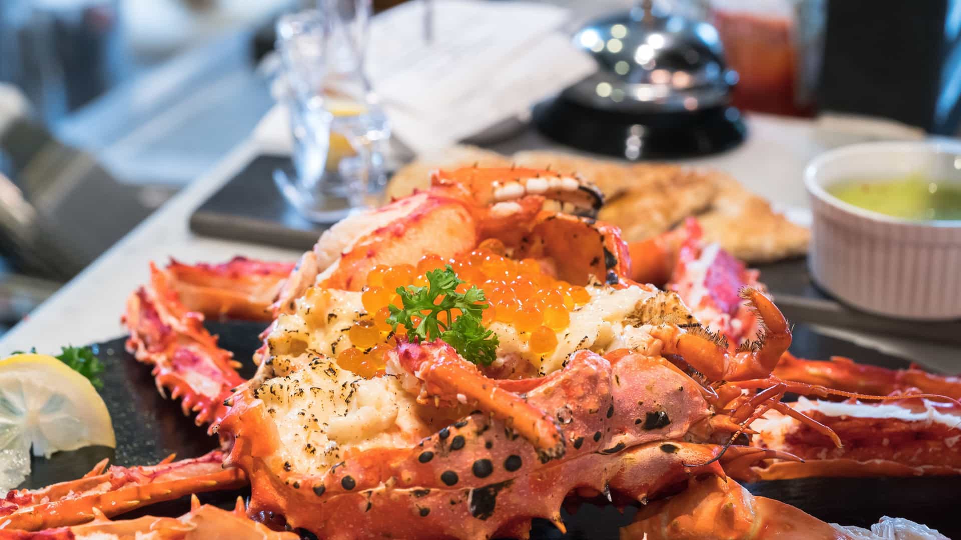 A close-up, top-down view of bright red, steamed King or Snow crab legs and claws piled on a dark plate. The crab is generously coated in a glistening, thick, red-orange spicy chili sauce and garnished with fresh green herbs.