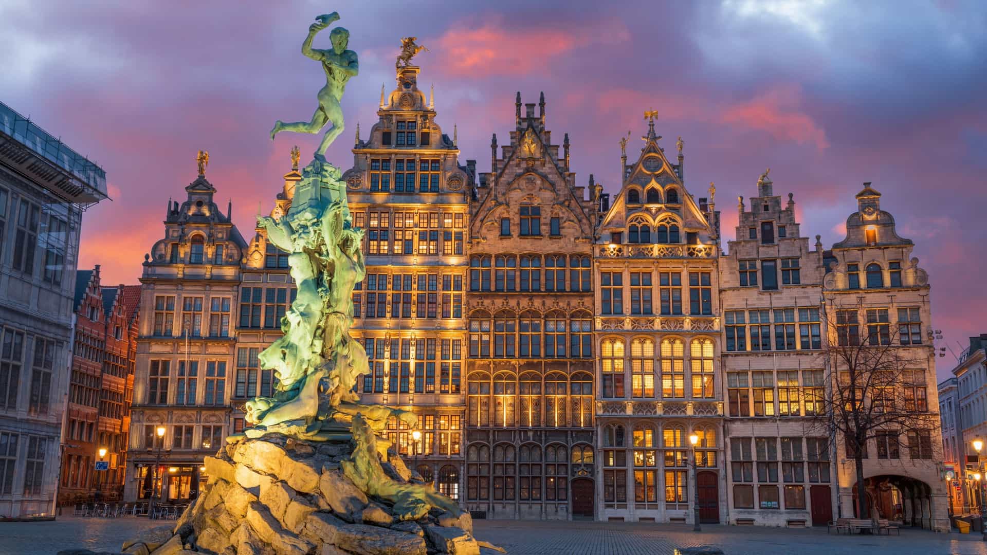 A horizontal photo of the Grote Markt in Antwerp, Belgium, at twilight. The central Brabo fountain and statue are visible in the foreground. The historic guild houses with their ornate, illuminated facades line the square, with the Antwerp City Hall standing out in the background. The sky is a deep blue, and the lights from the buildings and streetlamps are reflected on the wet pavement.