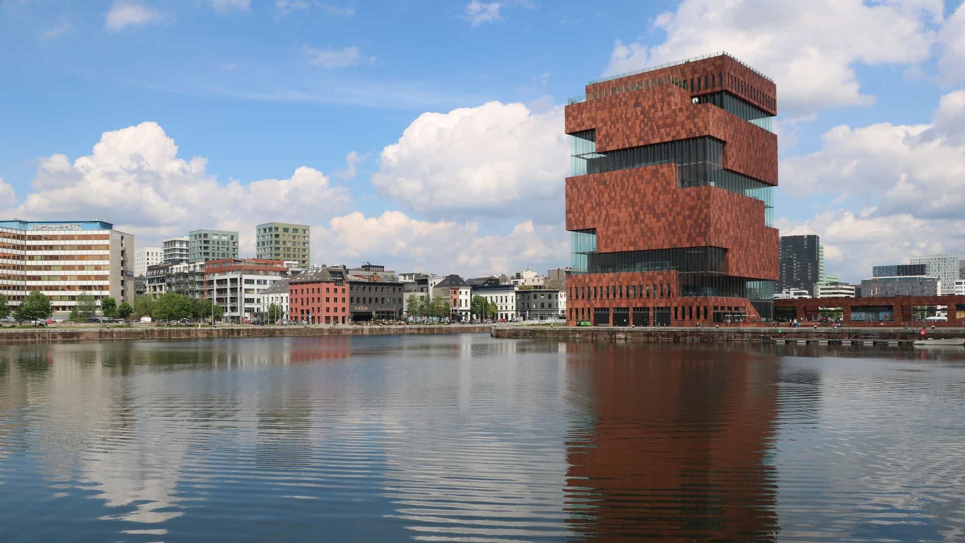 The MAS (Museum aan de Stroom) in Antwerp, Belgium. The modern, sixty-meter-high tower is composed of stacked blocks clad in red Indian sandstone. A spiral public gallery wraps around the exterior, enclosed by a distinctive, undulating curtain of curved glass panels, all set beside a harbor dock.