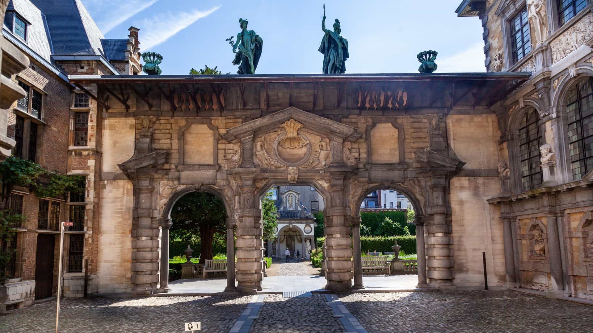 A view of the Italianate portico, designed as a triumphal arch, inside the Rubens House in Antwerp, Belgium. The structure features rusticated stone blocks, a central arched passage, and decorative niches that house classical bronze statues, such as Minerva and Mercury.