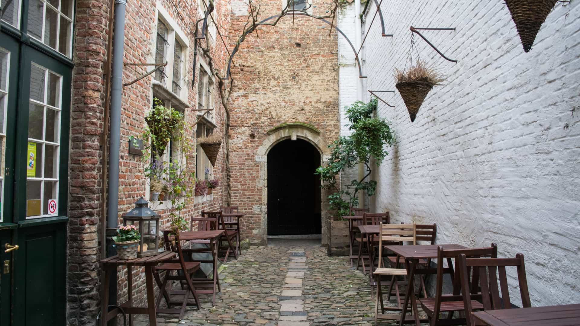 A narrow, historic cobblestone alleyway in the Vlaeykensgang, a hidden medieval quarter in the city center of Antwerp, Belgium. The scene is enclosed by old brick buildings with small windows and is characterized by its tranquil, time-worn atmosphere.