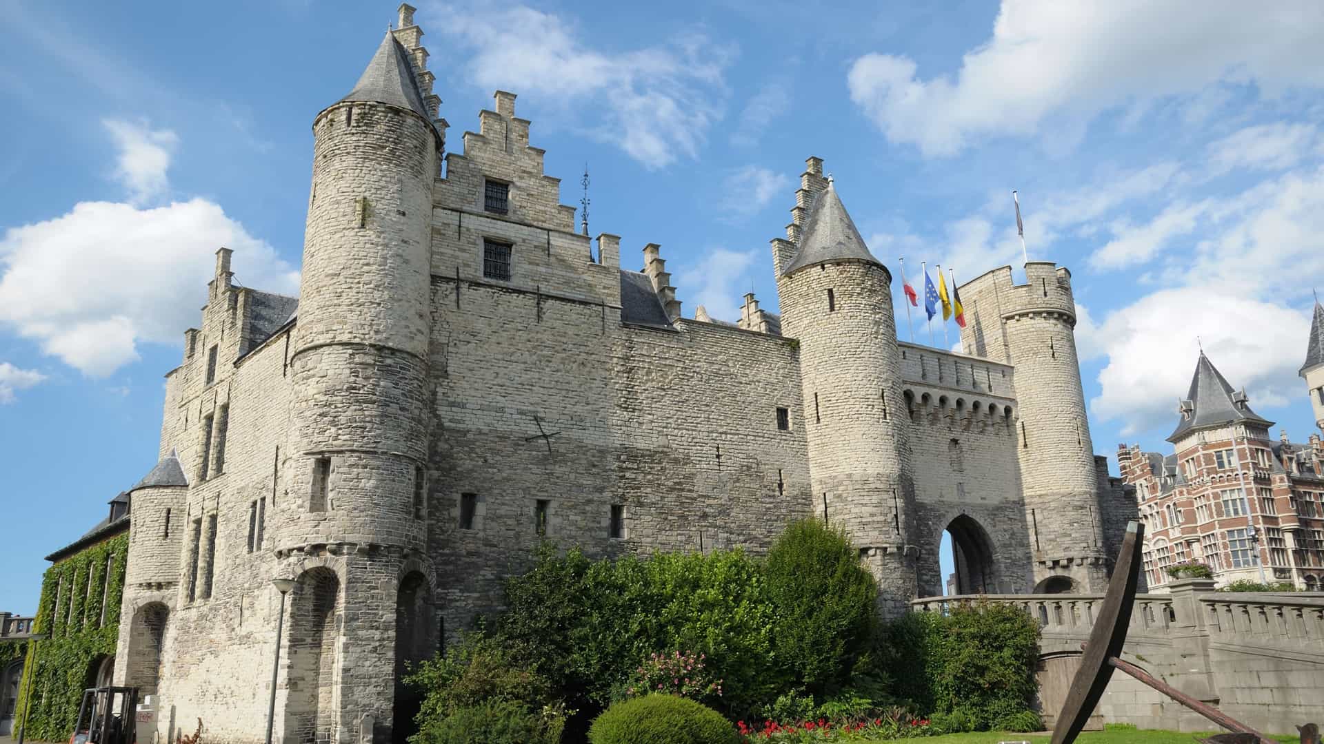 A horizontal photo of Het Steen, a medieval fortress in Antwerp, Belgium. The castle is situated on the banks of the Scheldt river. The stone building with its lighter and darker sections and an ornate gateway is clearly visible. The statue of the giant Lange Wapper stands on the bridge leading to the entrance. The photo is taken on a sunny day with a clear blue sky.