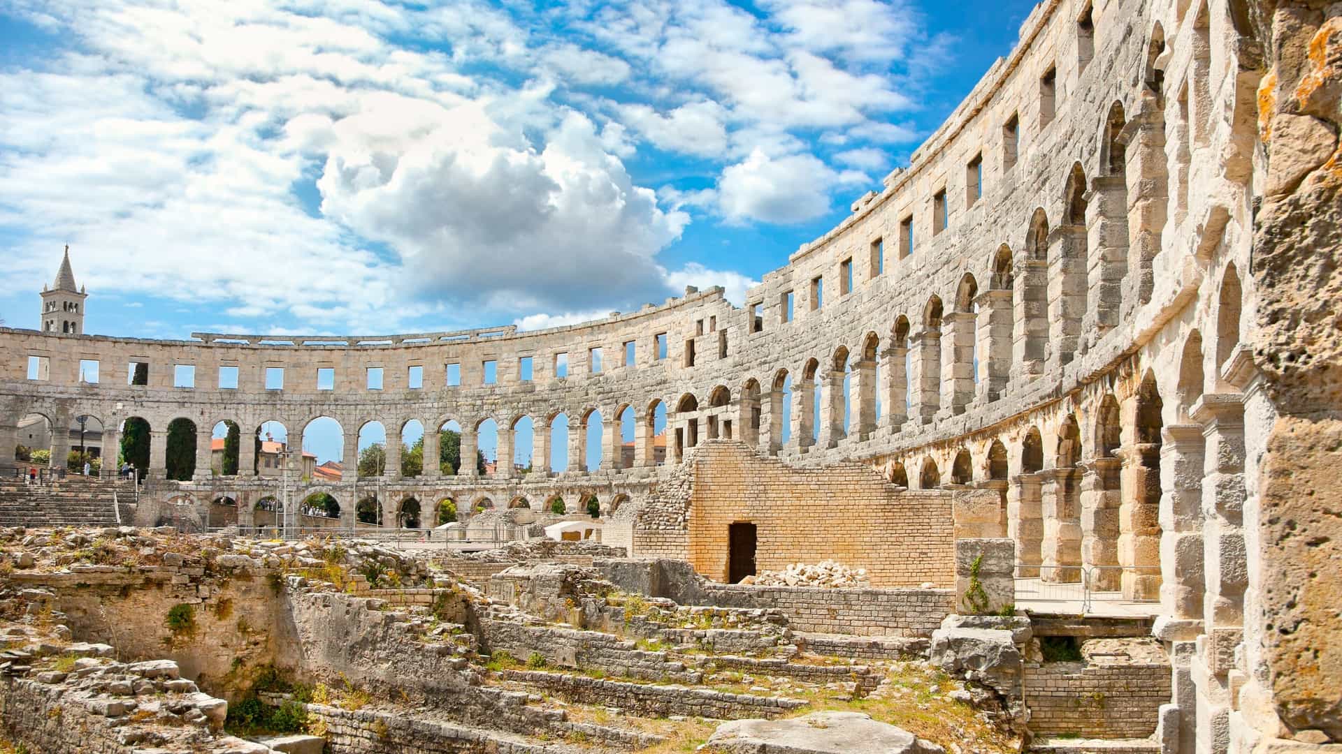 A horizontal photo of the Roman Amphitheater (Arena) in Pula, Croatia, on a sunny day. The ancient stone structure is shown from the outside, highlighting its well-preserved, multi-tiered arches and walls. The sky is clear, and the image captures the impressive scale and architectural detail of the historic landmark.