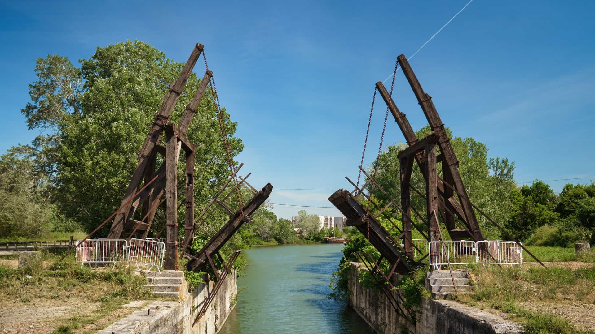 The historic Pont Van Gogh (also known as the Langlois Bridge), a reconstruction of the classic double-beam wooden drawbridge that spans the canal outside of Arles, France. The bridge, famous for being depicted in multiple paintings by Vincent van Gogh, is seen over calm water in a rural landscape under a clear sky.