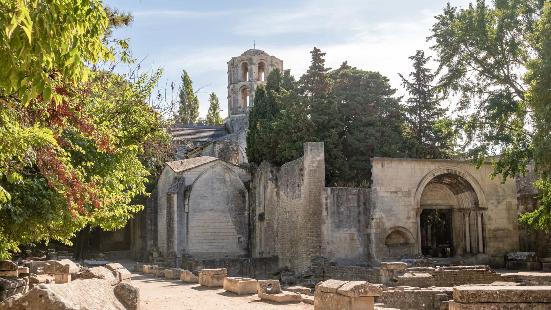 A view of Les Alyscamps, an ancient Roman necropolis in Arles, France. The image shows a shaded avenue lined with rows of ancient stone sarcophagi. The path is bordered by trees, creating a perspective that leads towards a Romanesque church in the distance. The scene conveys a sense of historical tranquility.