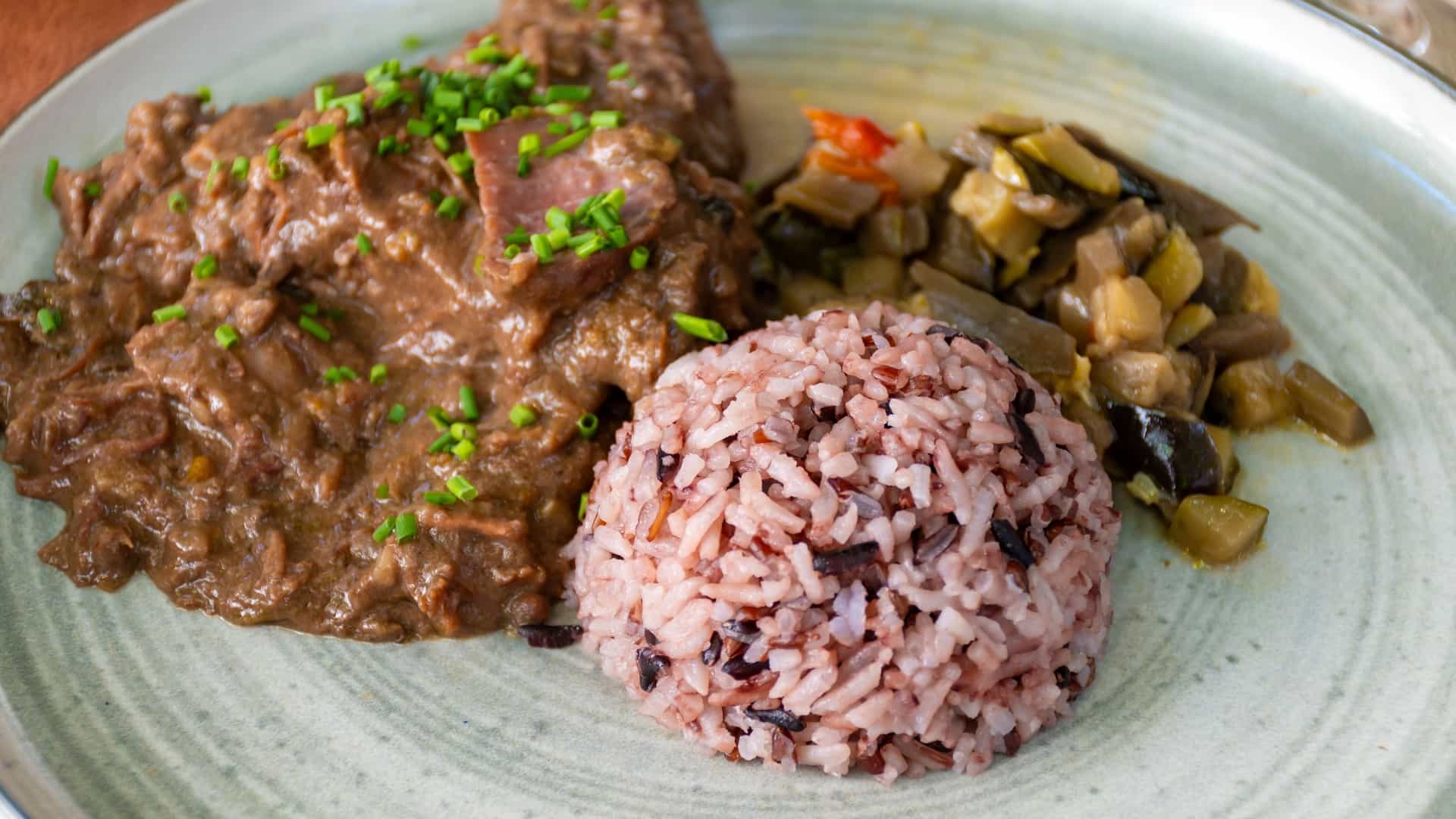 A close-up of a plate containing Gardiane de Taureau (or Daube Camarguaise), a traditional French stew from the Camargue region. The rich, dark red, slow-cooked bull or beef stew is served with a side of white Camargue rice and is garnished with fresh parsley.