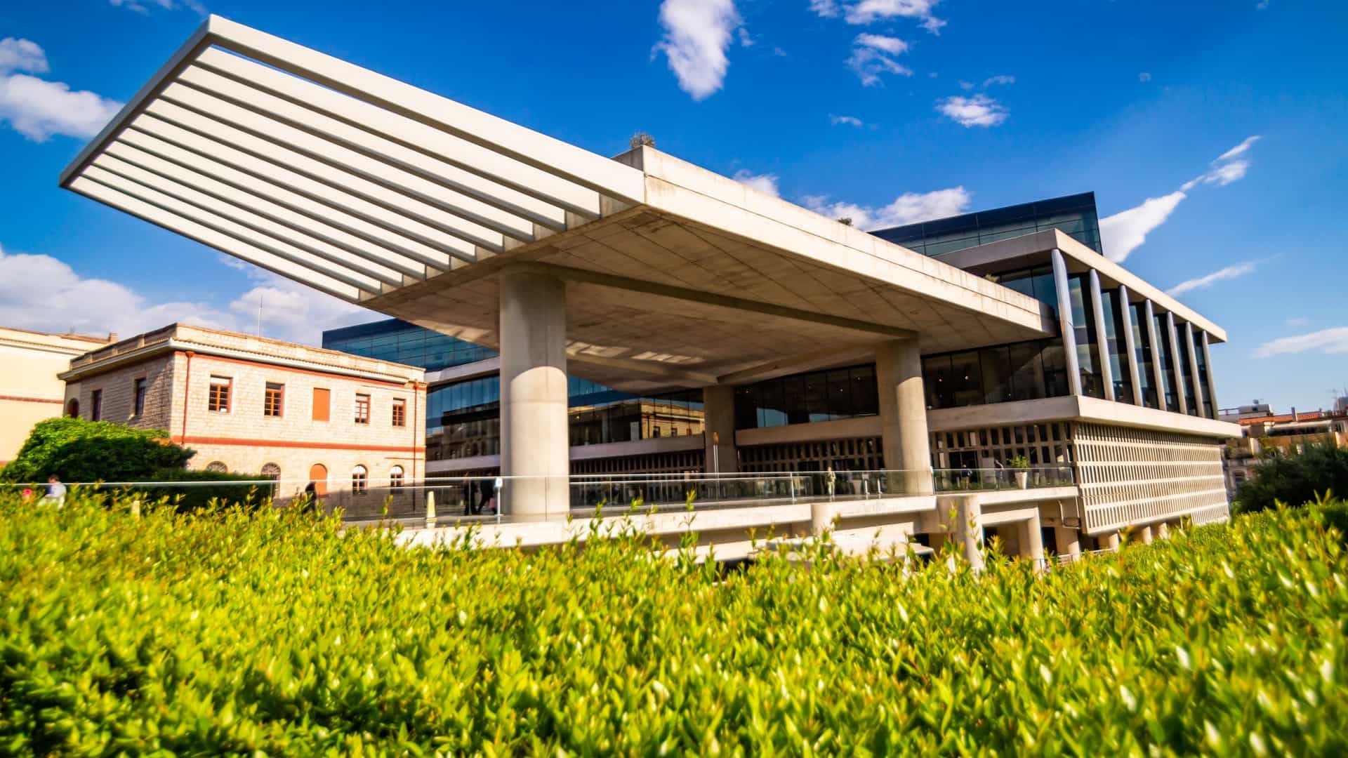 The modern New Acropolis Museum in Athens, Greece, designed by Bernard Tschumi Architects. The exterior view shows the building's distinctive layered structure, with the top, glass-enclosed Parthenon Gallery set askew to align with the ancient temple, resting on the middle section of raw concrete and columns.
