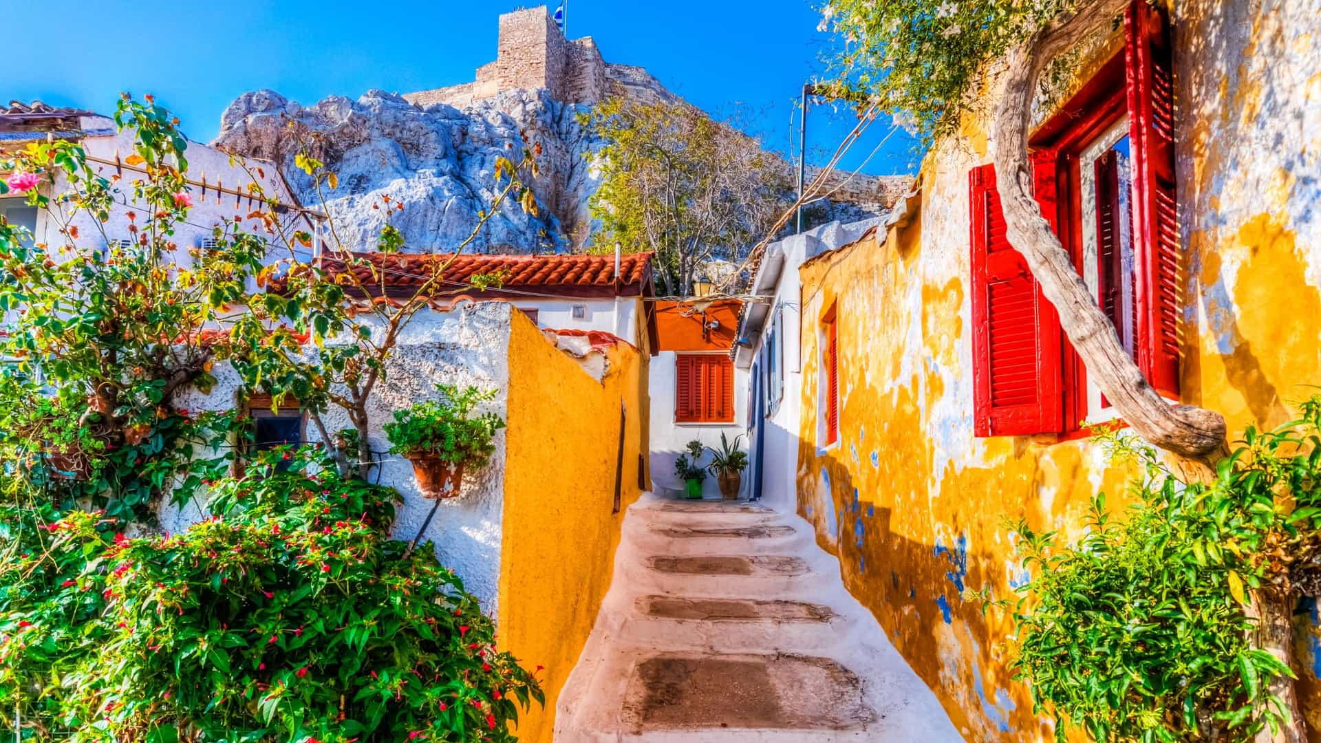 A view of a street in the Plaka district of Athens, Greece. The image shows a narrow, charming street with tables and chairs from a cafe, surrounded by traditional buildings. The street is lined with plants, and a sense of a bustling, historical neighborhood is conveyed.