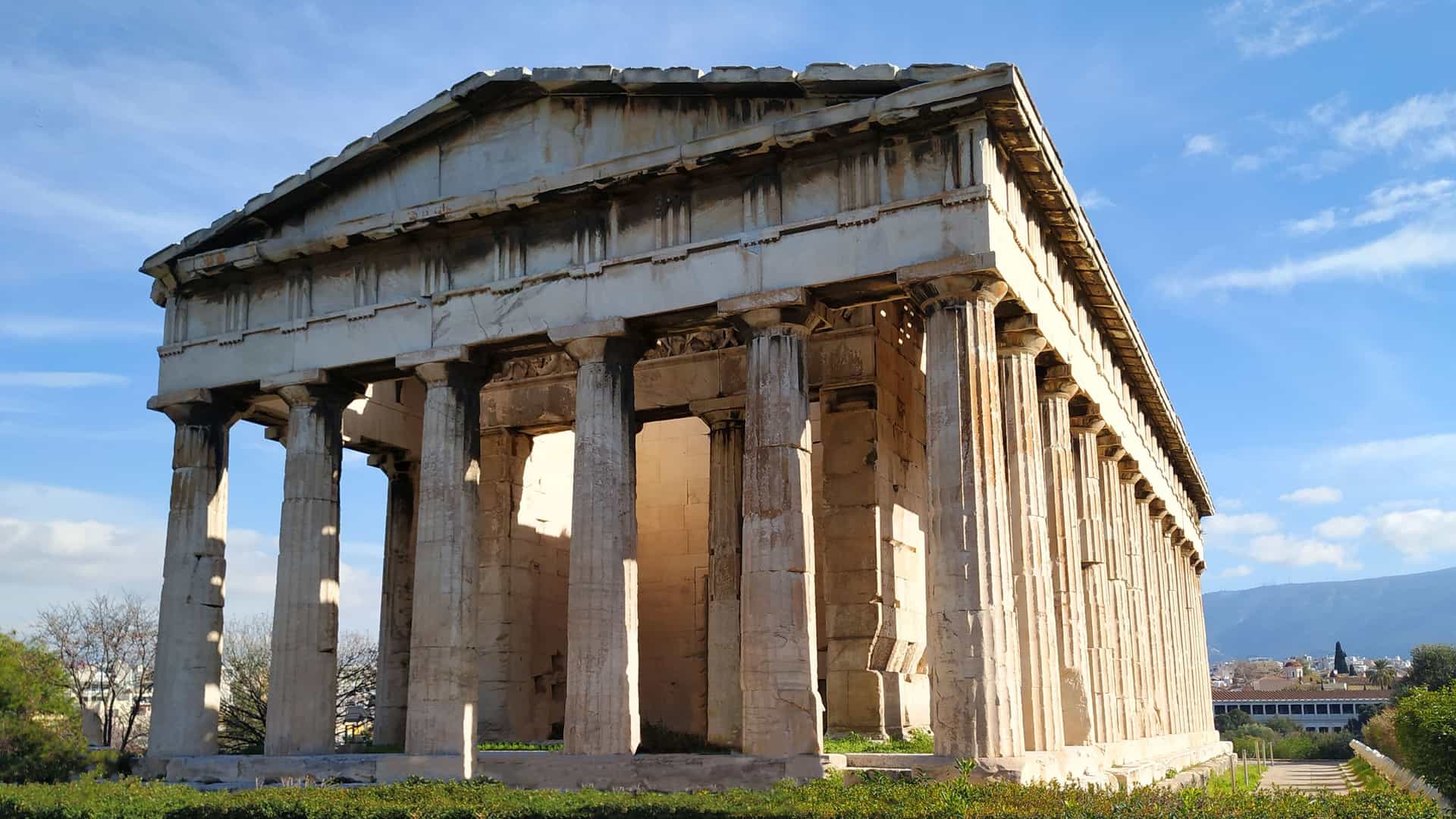 A view of the archaeological site of the Ancient Agora in Athens, Greece. The image features the well-preserved Temple of Hephaestus, a Doric temple with classical columns. The site includes other scattered ruins, pathways, and greenery, set against the backdrop of the city and the surrounding hills.