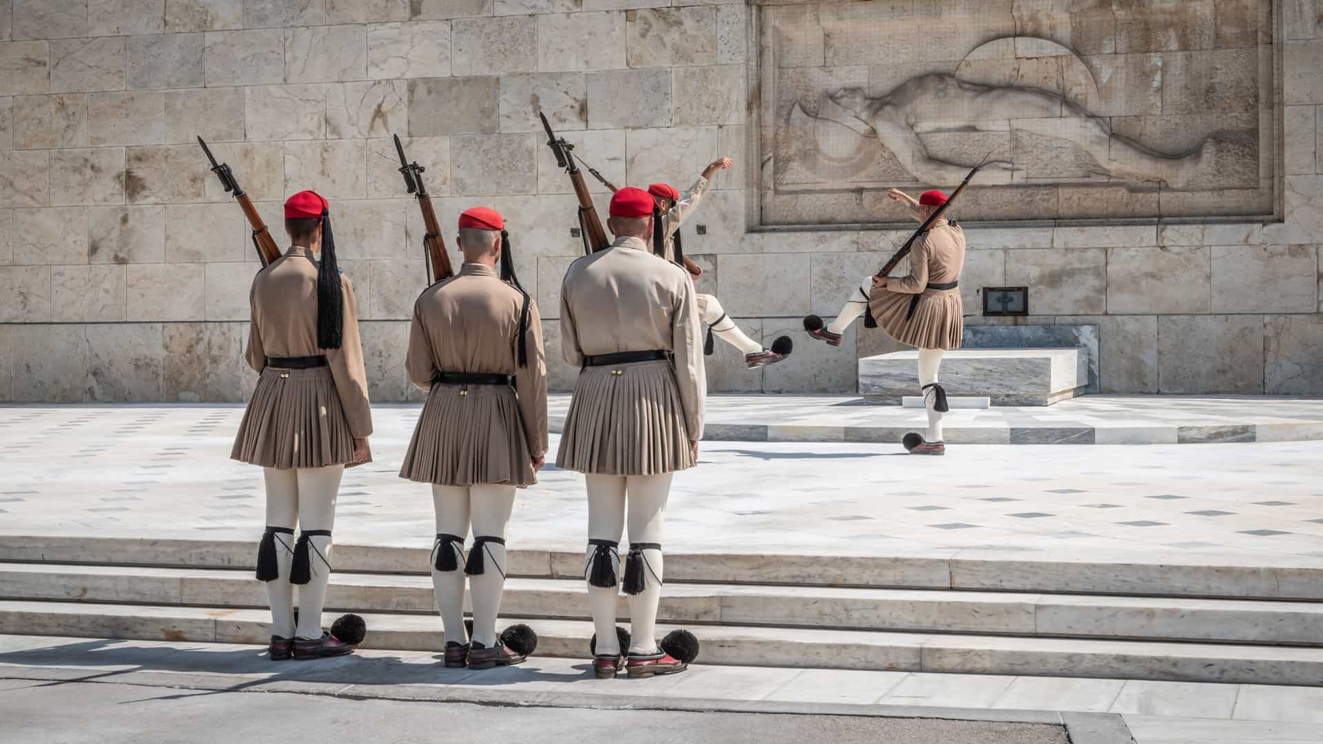 Two members of the Greek Presidential Guard, known as Evzones, standing guard in their traditional ceremonial uniforms. They are positioned in front of the Tomb of the Unknown Soldier in Athens, Greece, with the Hellenic Parliament building visible in the background. The guards are wearing their pleated kilts, red caps, and shoes with black pompoms.