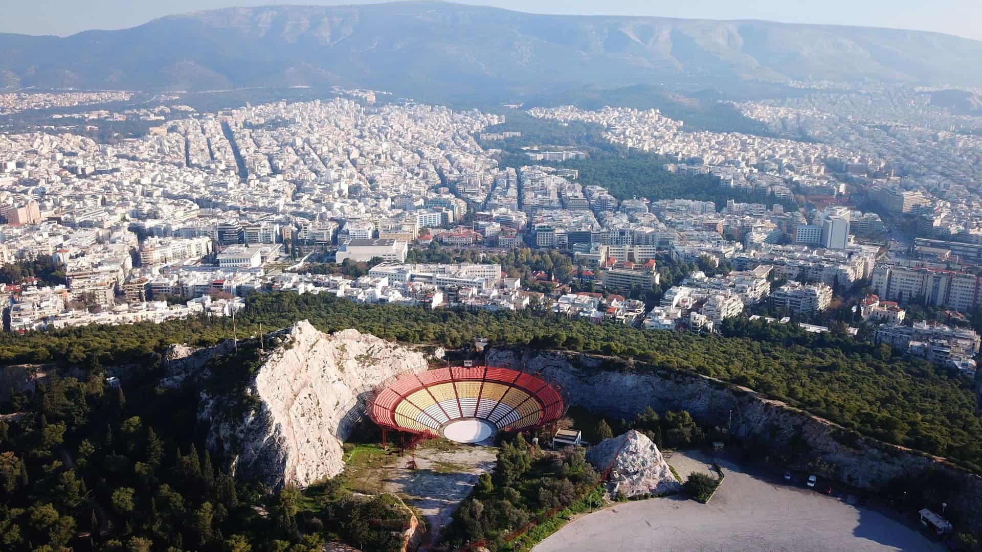 An aerial drone photo of the Lycabettus Theatre, an open-air amphitheater with tiered seating carved into the slopes of Lycabettus Hill in Athens, Greece. The theater's circular stage and seating are visible, with the urban landscape of the city sprawling out below it.
