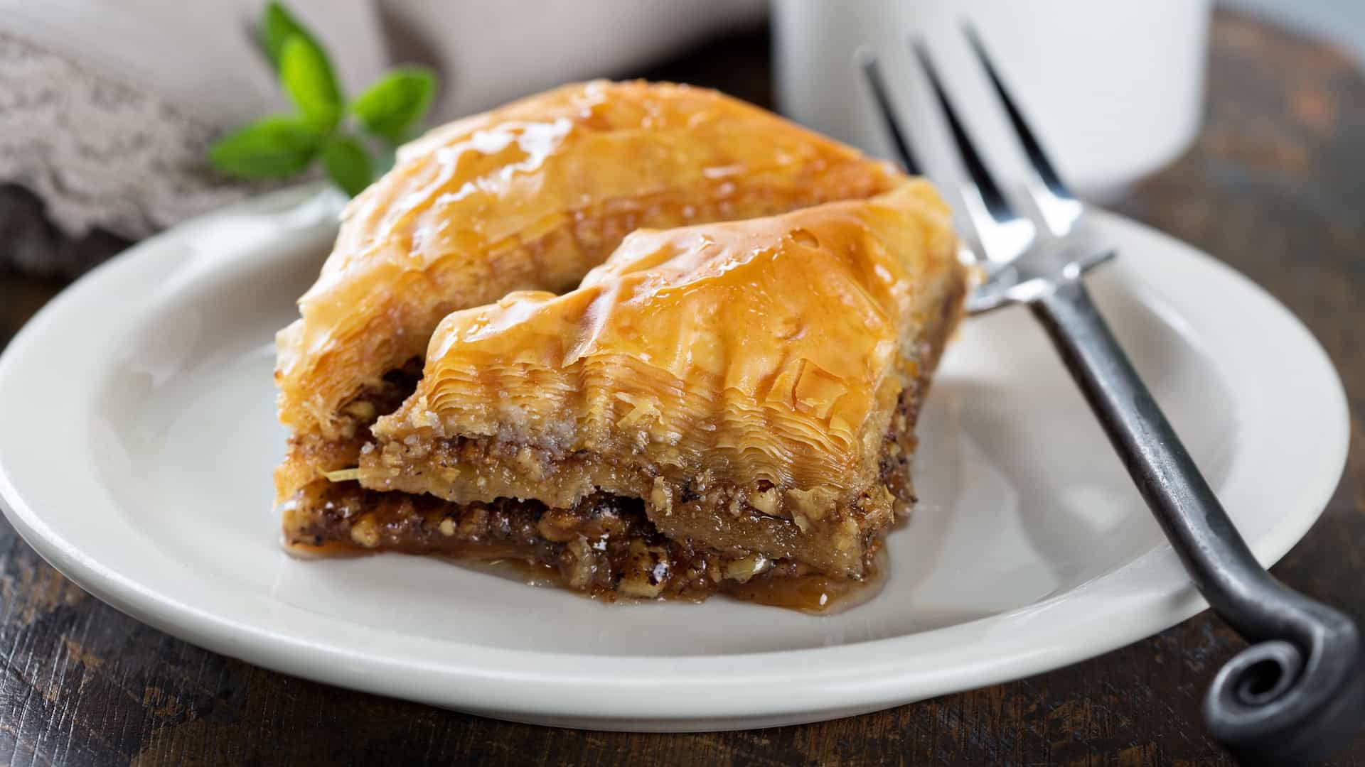 A close-up, tabletop view of a piece of baklava, a traditional Middle Eastern and Turkish pastry, on a small dessert plate. The pastry is made of layers of phyllo dough, nuts, and syrup. A small cup of coffee is next to the plate.