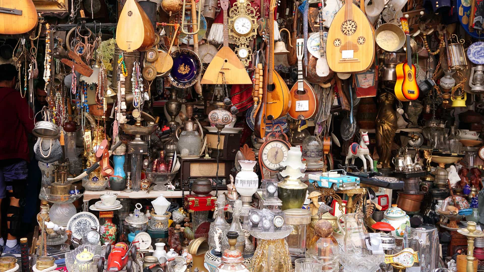 A view of a small, packed antique shop on a street in the Monastiraki Flea Market in Athens, Greece. The shop entrance and open facade are overflowing with various unique items, including several musical string instruments (likely bouzoukis or guitars) hanging from the ceiling, old wooden furniture, metal objects, ceramics, and miscellaneous vintage items.